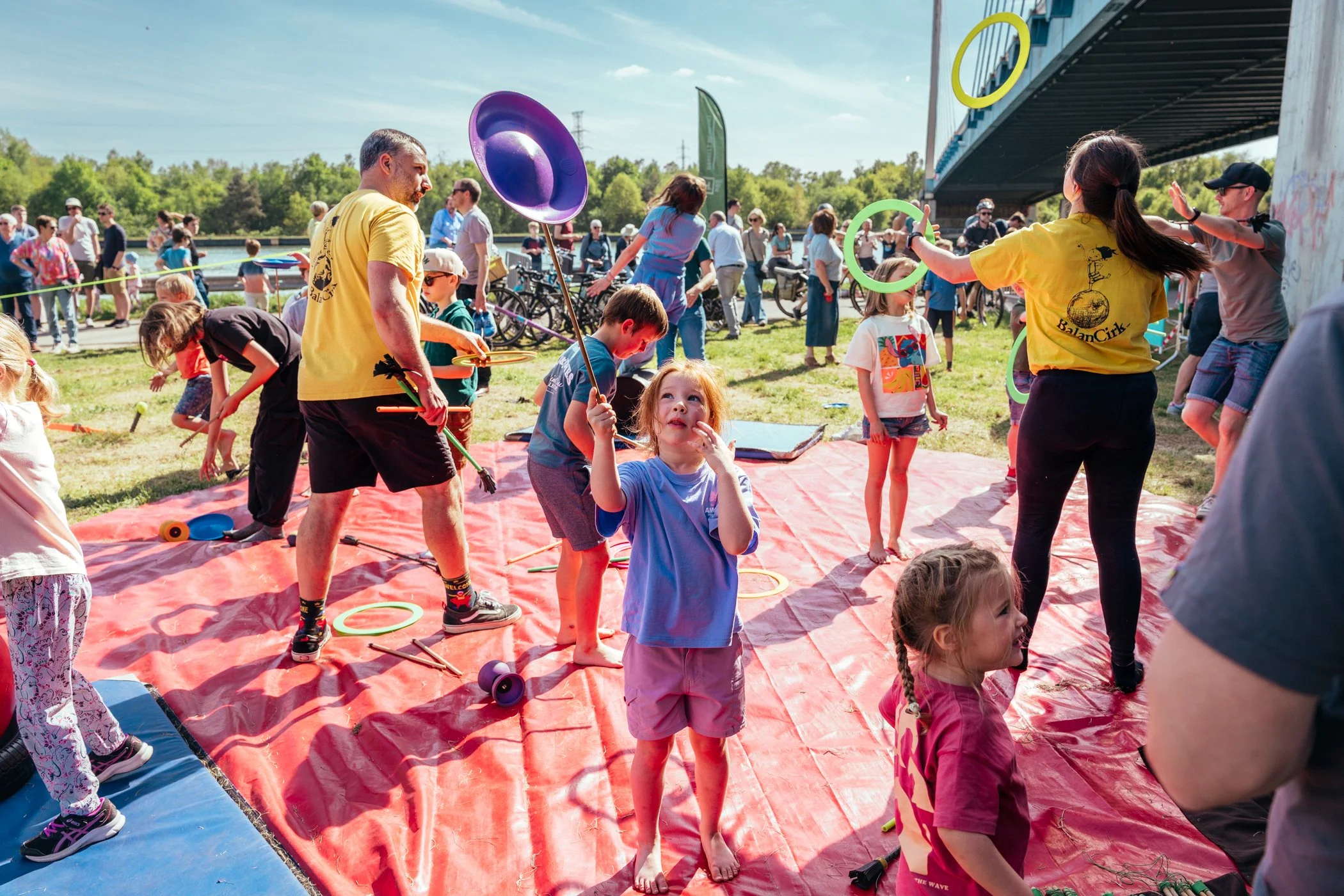 Kinderen en volwassenen die op een rode mat onder een brug acrobatie en jongleerkunstjes doen tijdens een outdoor festival.