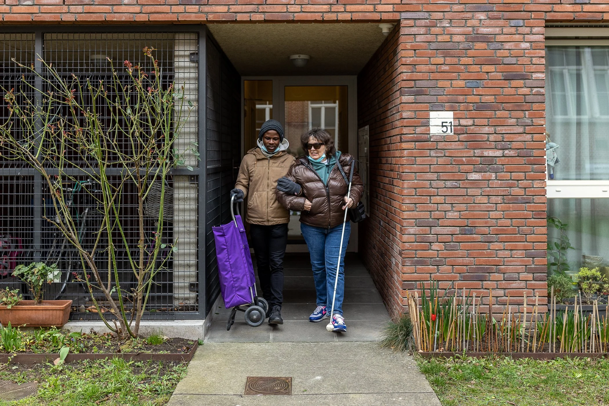 Twee vrouwen lopen samen uit een appartementencomplex, één met een zoomrol en de ander met een blindenshield.