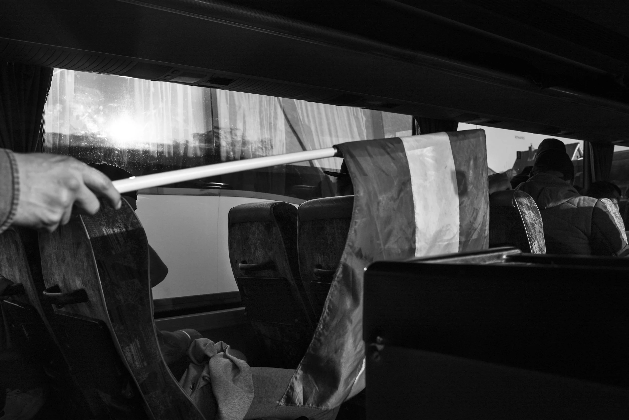 Binnenkant van een bus met een hand die een ruitenwissers vasthoudt. Mensen zitten op stoelen en kijken naar buiten, met zonlicht dat door de ramen schijnt.