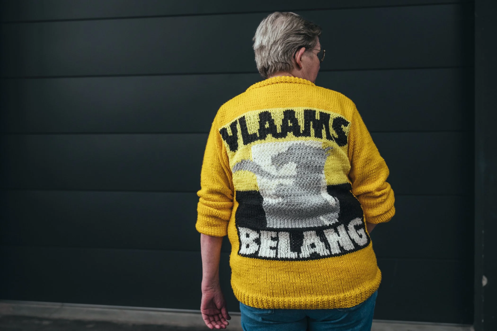 Persoon met grijs haar die een gele sweater draagt met de tekst 'YILLAHS BELANG' en een grafisch ontwerp aan de achterkant, staand voor een zwarte muur.