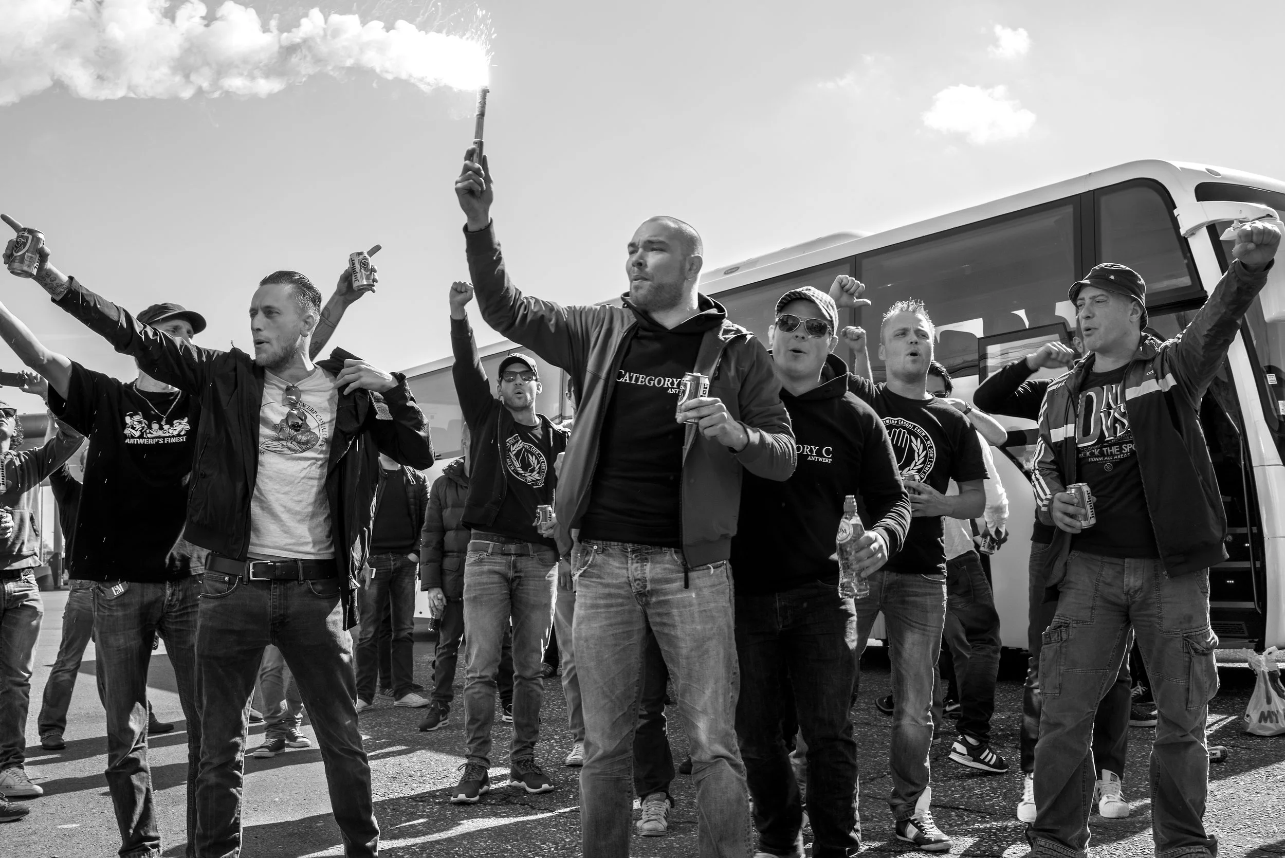 Een groep mannen die op straat samenkomen, sommige met armen omhoog, vlaggen en dranken in de hand, bij een bus, tijdens overdag.