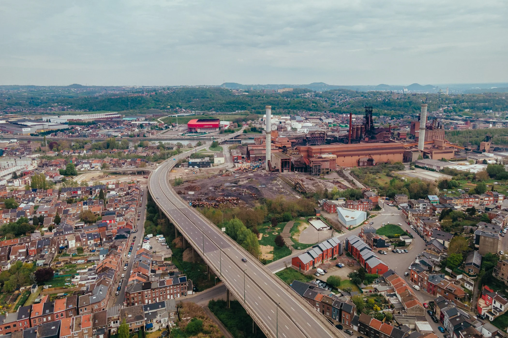 Luchtfoto van een stad met een grote fabriek, wegen en woonwijken omgeven door groene heuvels en landschap.
