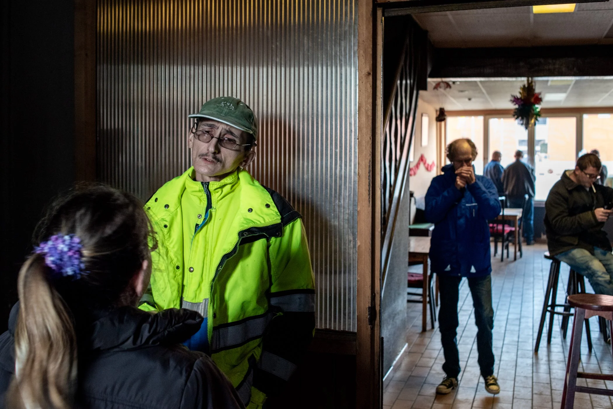Man in een felgele jas en groen pet heeft een gesprek met een vrouw met een paardenstaart in een café.