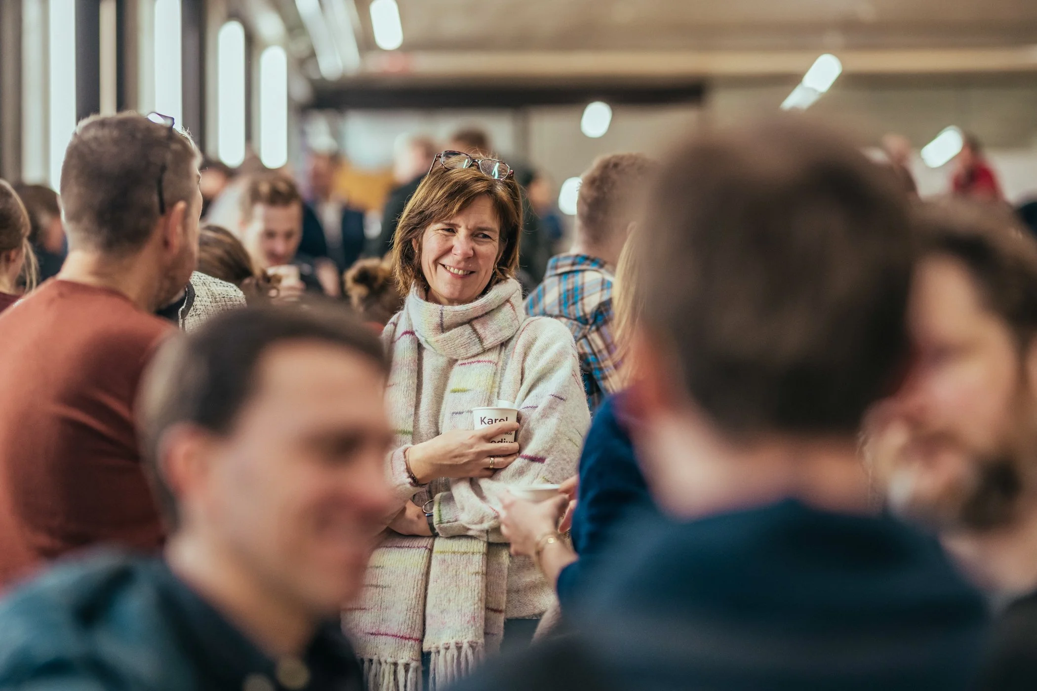 Vrouw met korte bruin haar, driekwart profiel, glimlachend, in een grote groep mensen binnen, met een lichte trui en een scarf, tijdens een sociale bijeenkomst.