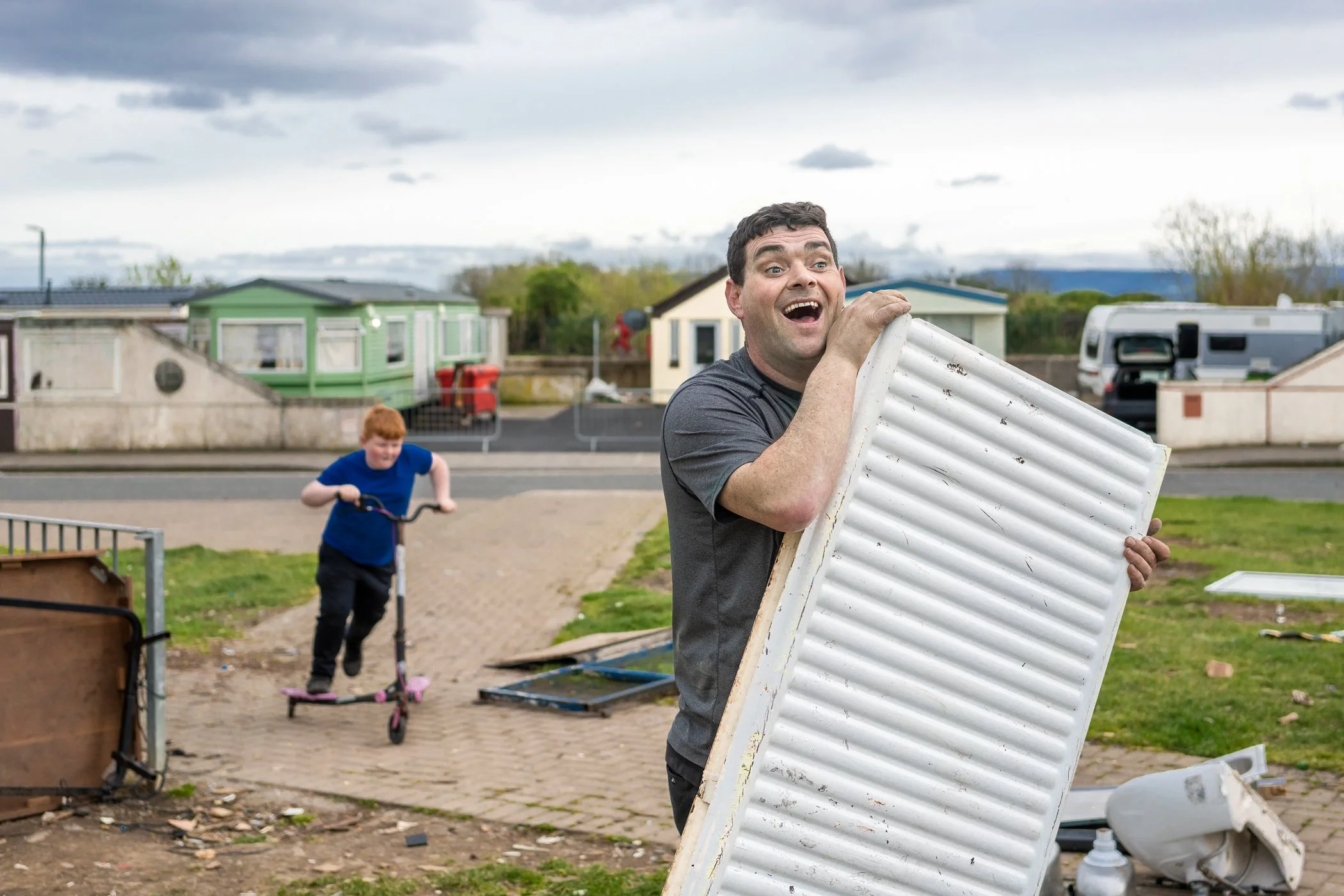 Man die een verouderde radiator vasthoudt terwijl een jongen op een scooter achter hem rijdt, op een rommelige buitenruimte met woningen op de achtergrond.