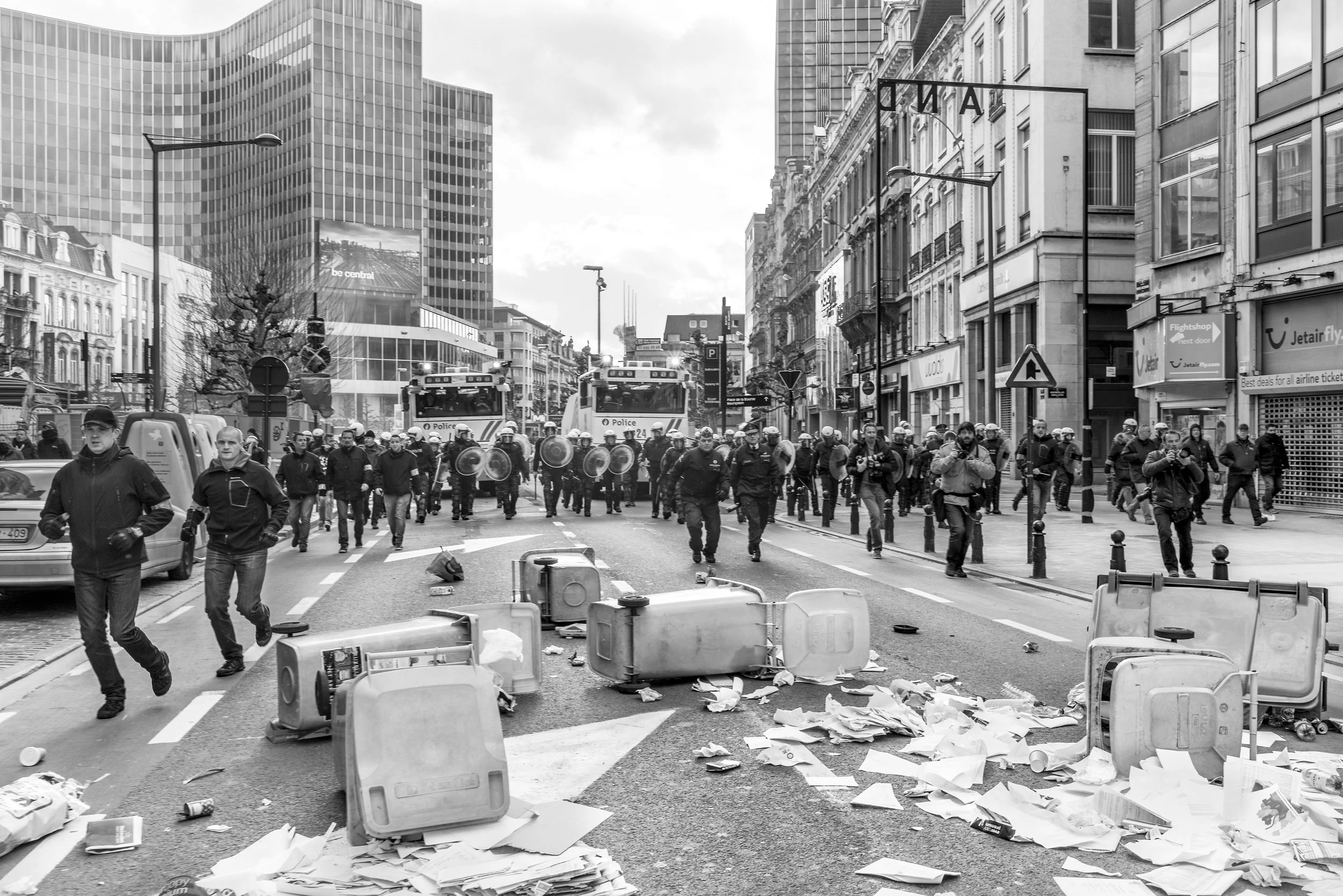 Straat met op de voorgrond rommel, afval en omgevallen vuilnisbakken. Maar daarachter politiemensen, politievoertuigen en een grote menigte, mogelijk demonstranten of betrokken bij een protest, op de straat.
