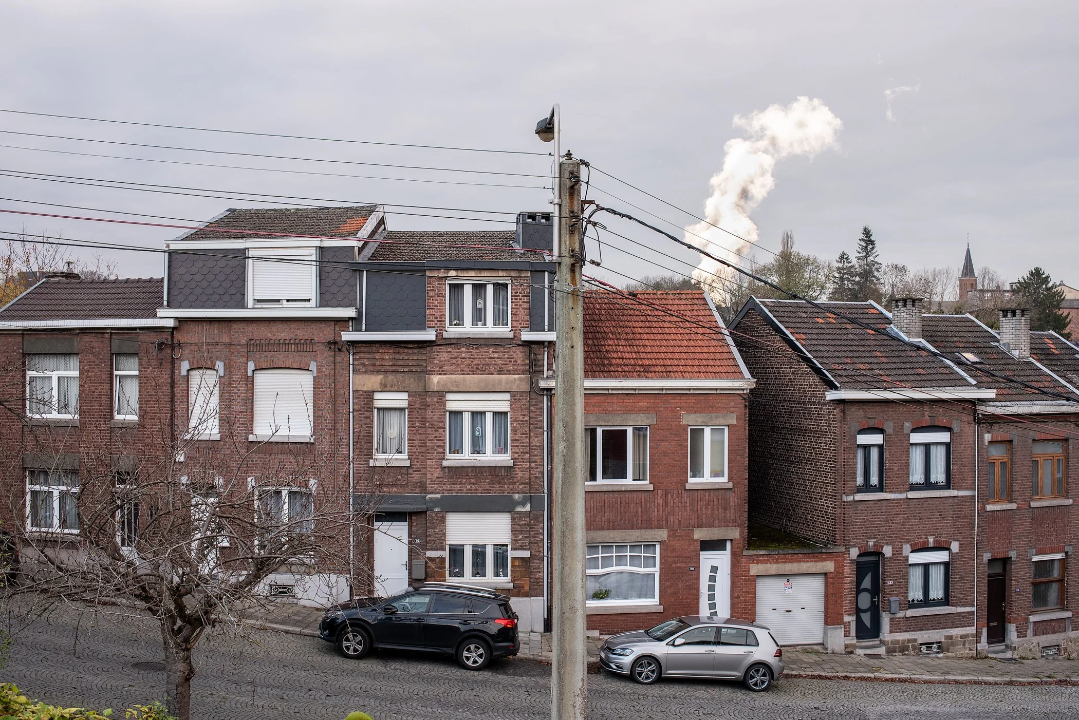 Rijen met oude en nieuwe bakstenen huizen, één met een extra verdieping en zonnepanelen op de daken, auto's geparkeerd voor de huizen, een boom zonder bladeren, elektriciteitslijnen en een groen uitzicht met bomen en een kerktoren in de achtergrond.