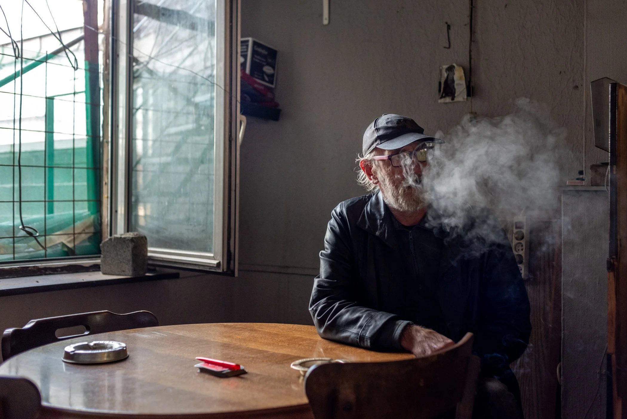 Een oudere man met een zwarte hoed, bril en een leren jas zit aan een houten tafel in een kamertje en blaast rook uit, waarschijnlijk van een sigaret of e-sigaret.