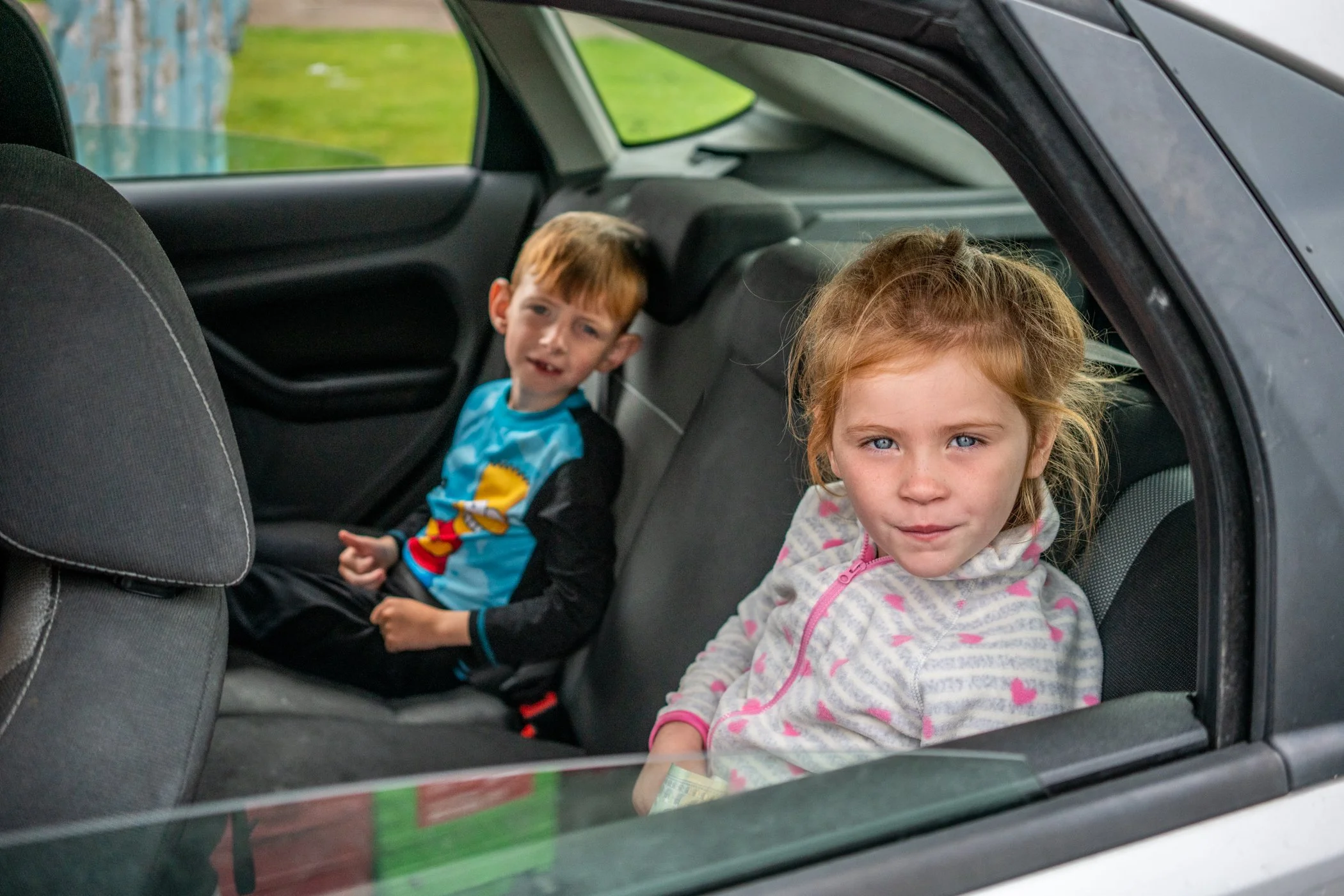 Twee kinderen zitten in de achterbank van een auto, de jongen met rood haar in een blauwe trui en het meisje met lichtbruin haar in een roze en grijze jas.