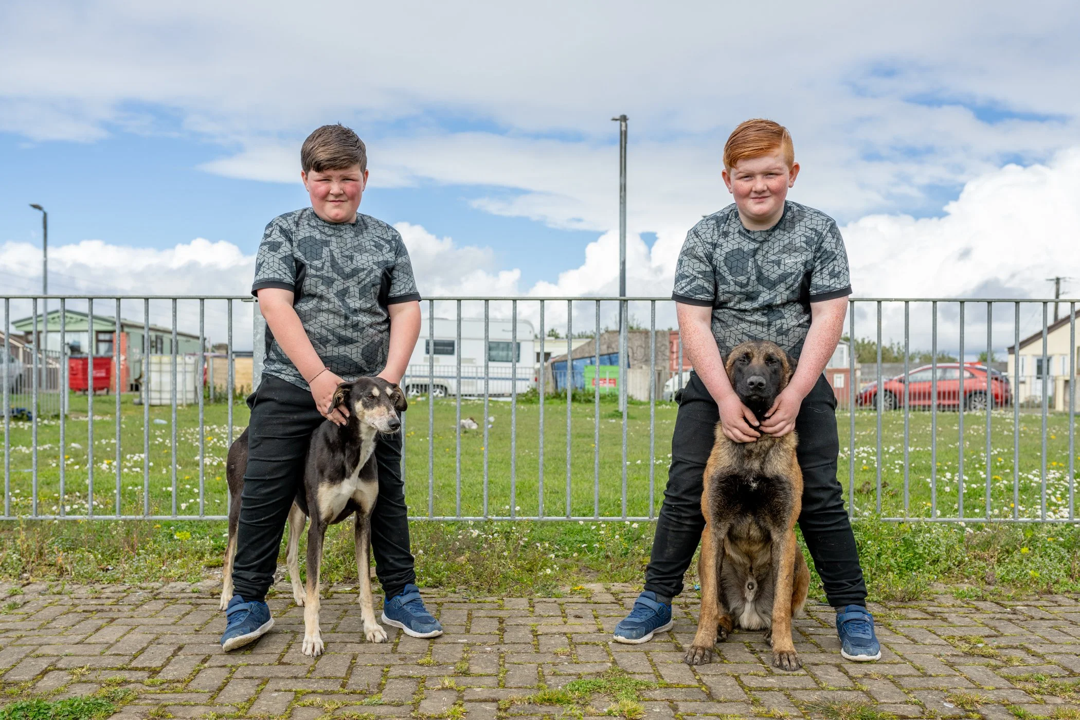 Twee jongens staan buiten op een stenen pad, elk met een hond voor hen. De jongens dragen vergelijkbare shirts en donker geklede broeken. De achtergrond toont een hek, grasveld, enkele gebouwen en een blauwe hemel met wolken.