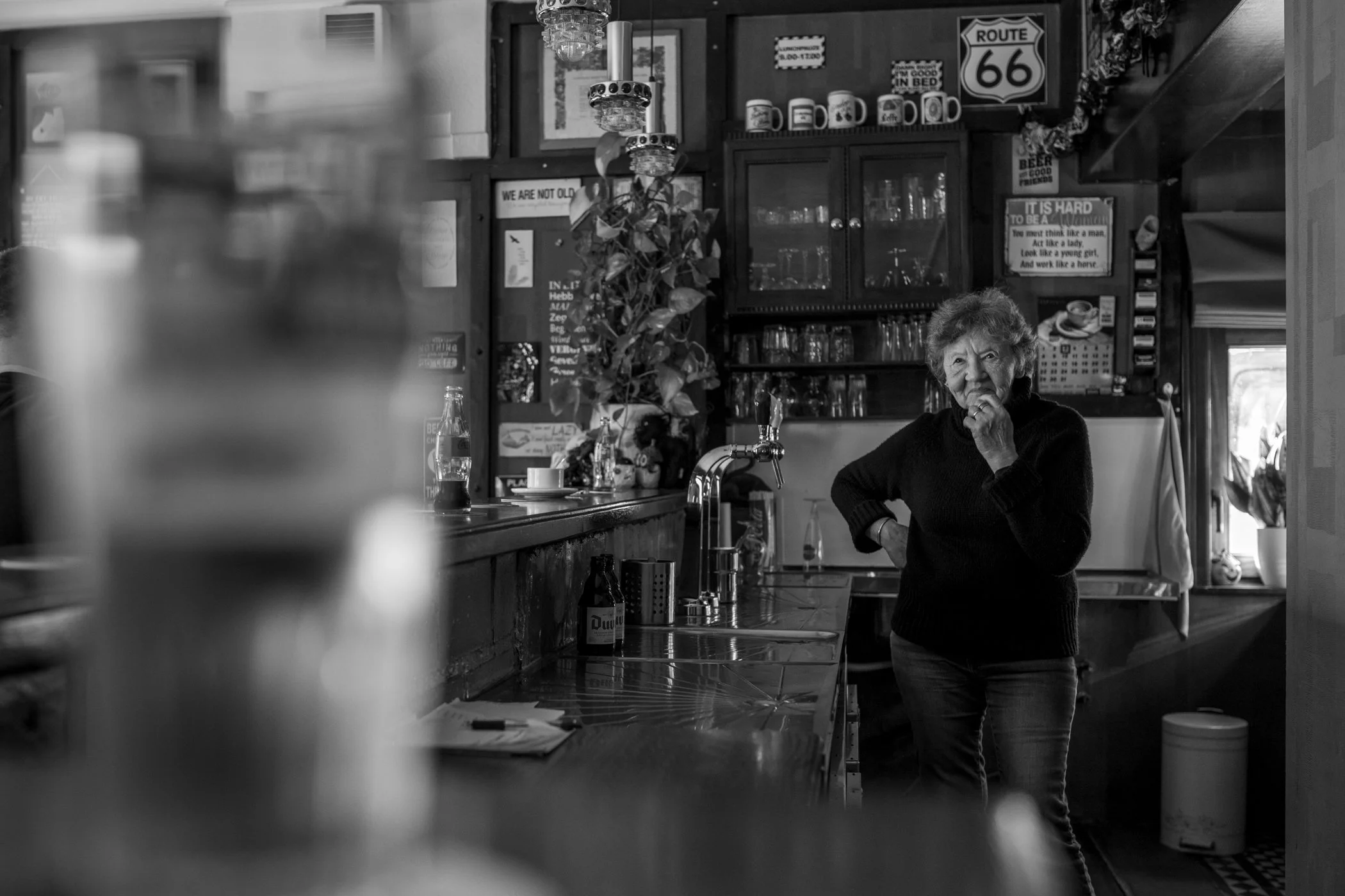 Een oudere vrouw staat in een restaurant of café, uitdrukking van gedachten of overpeinzing. De ruimte is ingericht met houten kasten, decoraties en posters aan de muur, en er is een gootsteen en keukengerei zichtbaar.