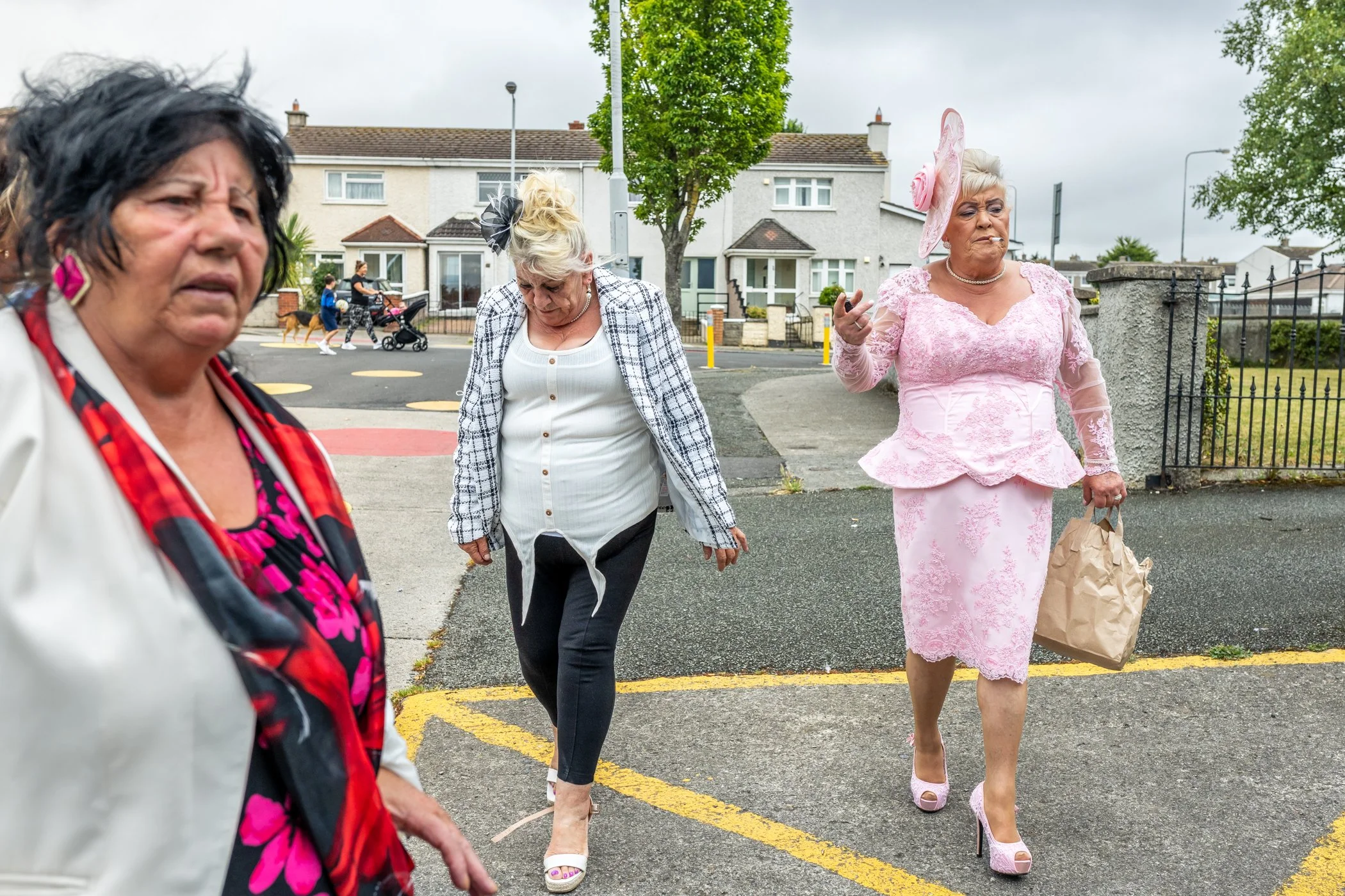Drie oudere dames wandelen op een straat in een woonwijk, één in een roze kledingstijl met hoed, een in een witte en blauwe outfit, en de derde in een witte jas met een zwart-roze jurk.