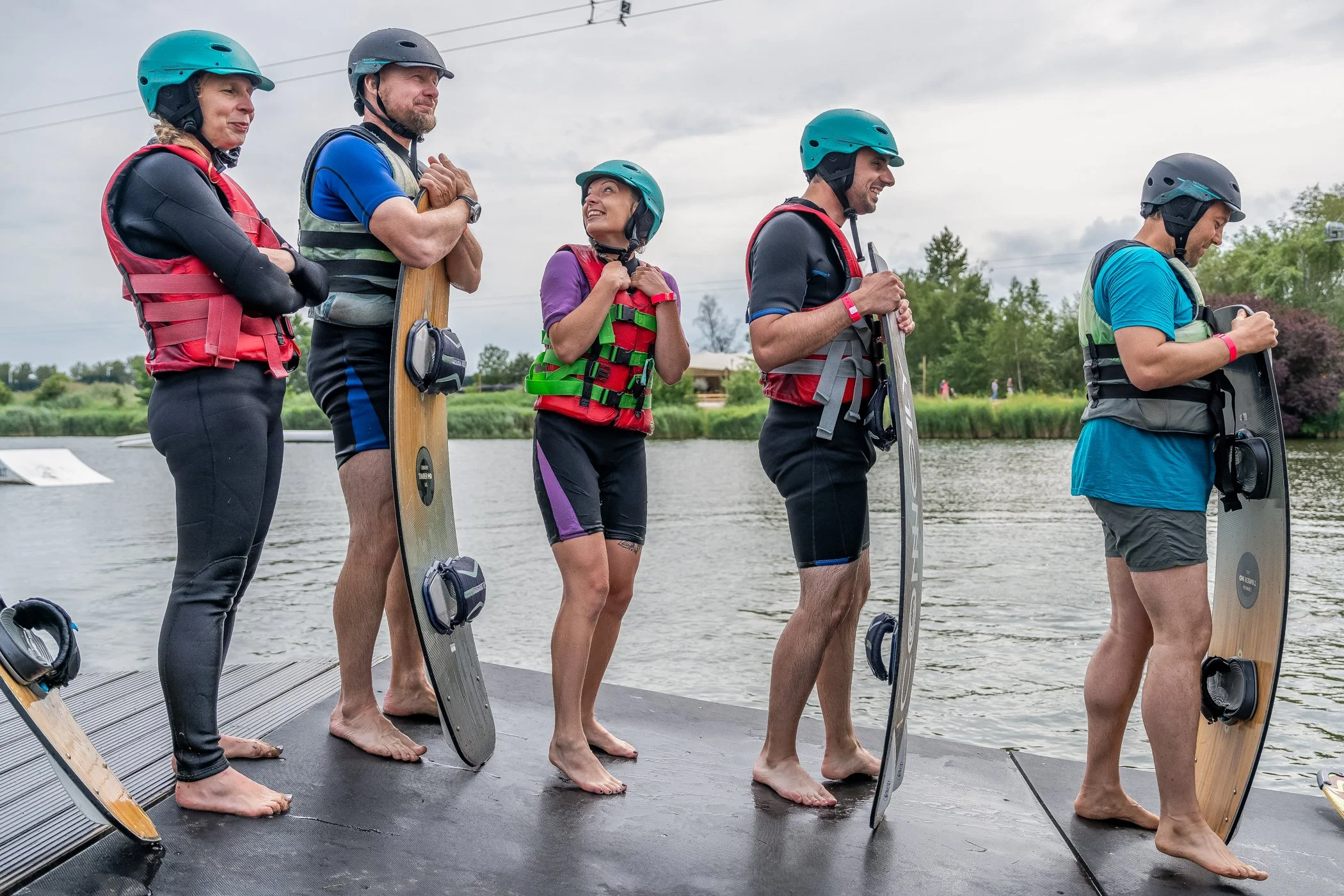 Vijf mensen met helmen en reddingsvesten staan op een boot, klaar om te gaan watersporten, met water en groen in de achtergrond.