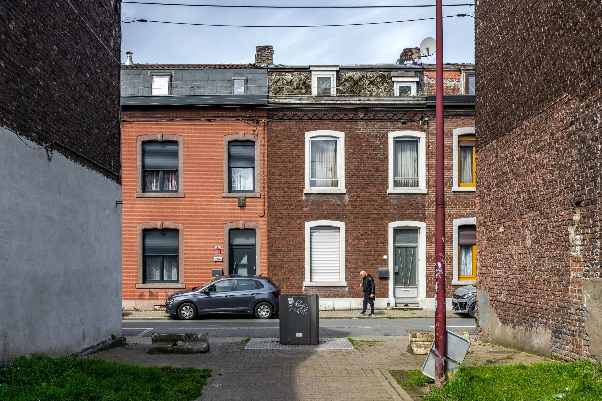 Straatbeeld met drie rijtjeshuizen, twee auto's geparkeerd, een man loopt langs, er is een elektriciteitskast en een verlichte lantaarnpaal, met een trottoir en muur in de voorgrond.