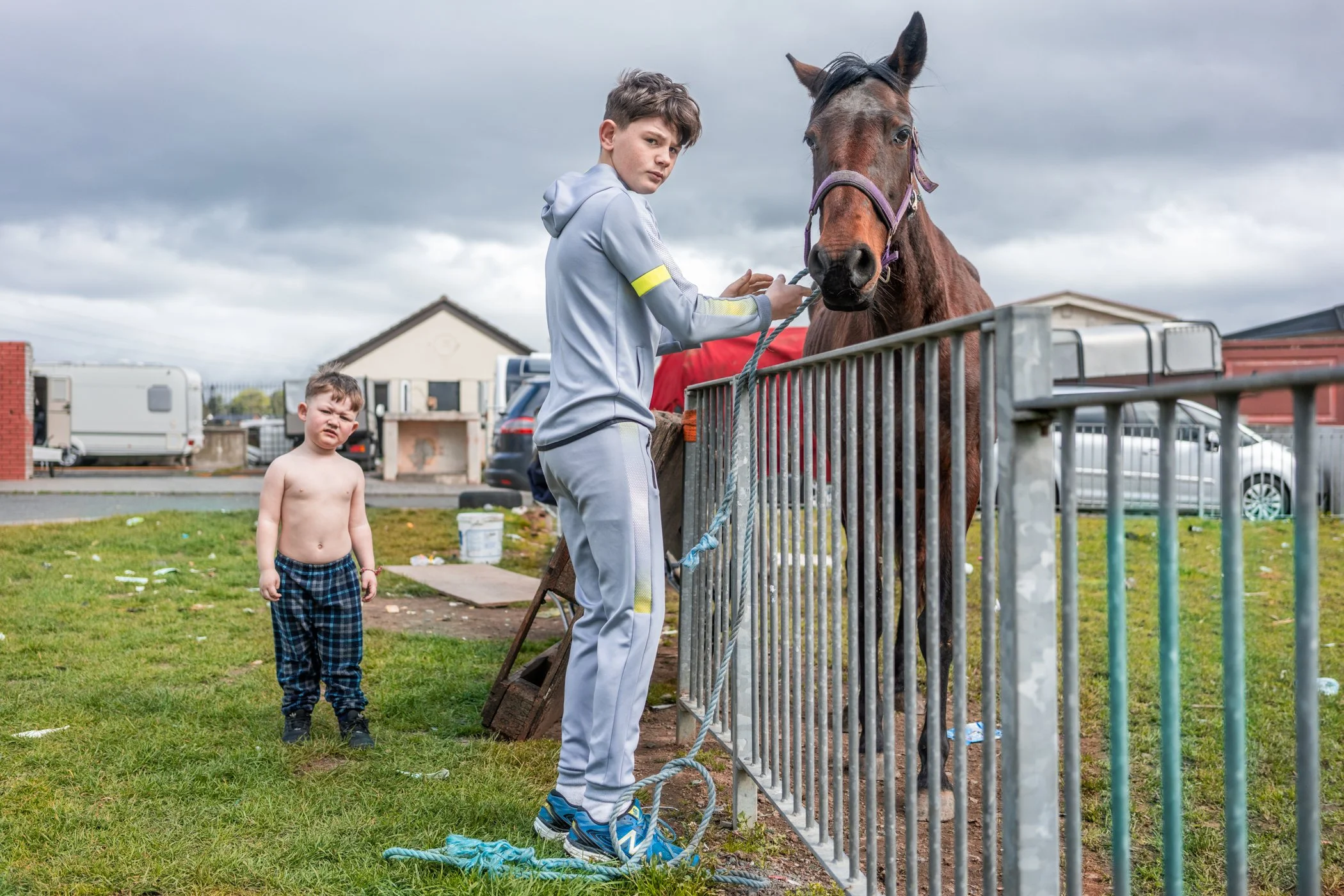 Een jonge jongen in een grijs trainingspak houdt een paard vast achter een metalen hek, terwijl een kleine jongen zonder shirt naar de camera kijkt op een grasveld bij een huis en auto's in de achtergrond.