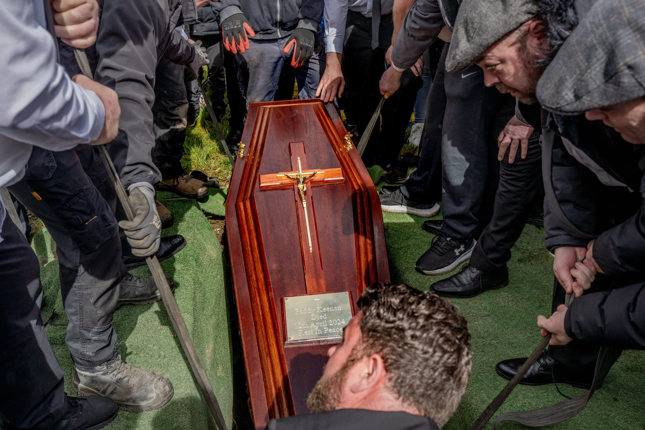 Mensen bij een begrafenis die rondom een gesloten houten kist staan, met een kruis en een grafetiket met de tekst 'Paddy Keenan Died 12 April 2024 Rest In Peace'.