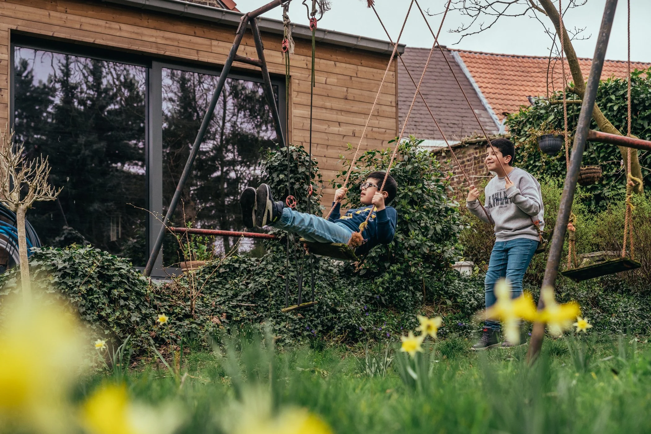 Twee jongens spelen op een schommel en een schommelstoel in een tuin met bloemen, planten en een huis met houten gevel en ramen.