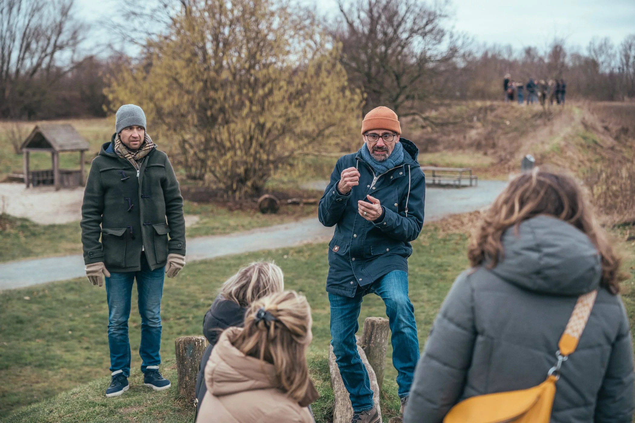 Mensen die in een park of natuurgebied bij koud weer gesprekken voeren, met vier mensen zittend en twee staand, in de achtergrond bladeren aan bomen en een klein water, met een groep mensen op de heuvel in de verte.