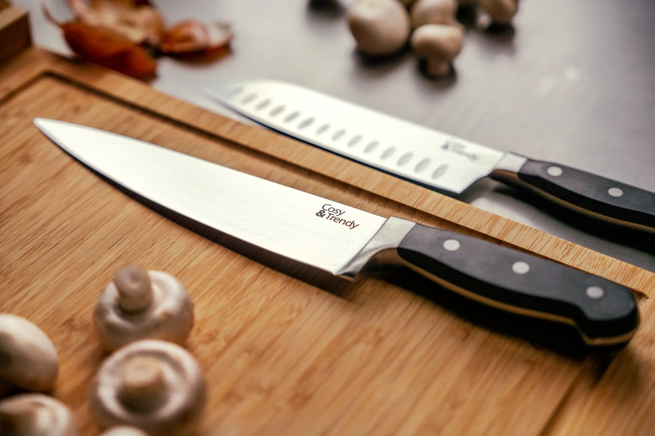 Keukenmes met zwarte handgreep en houten snede op houten snijplank, met witte champignons en ui op de voorgrond.
