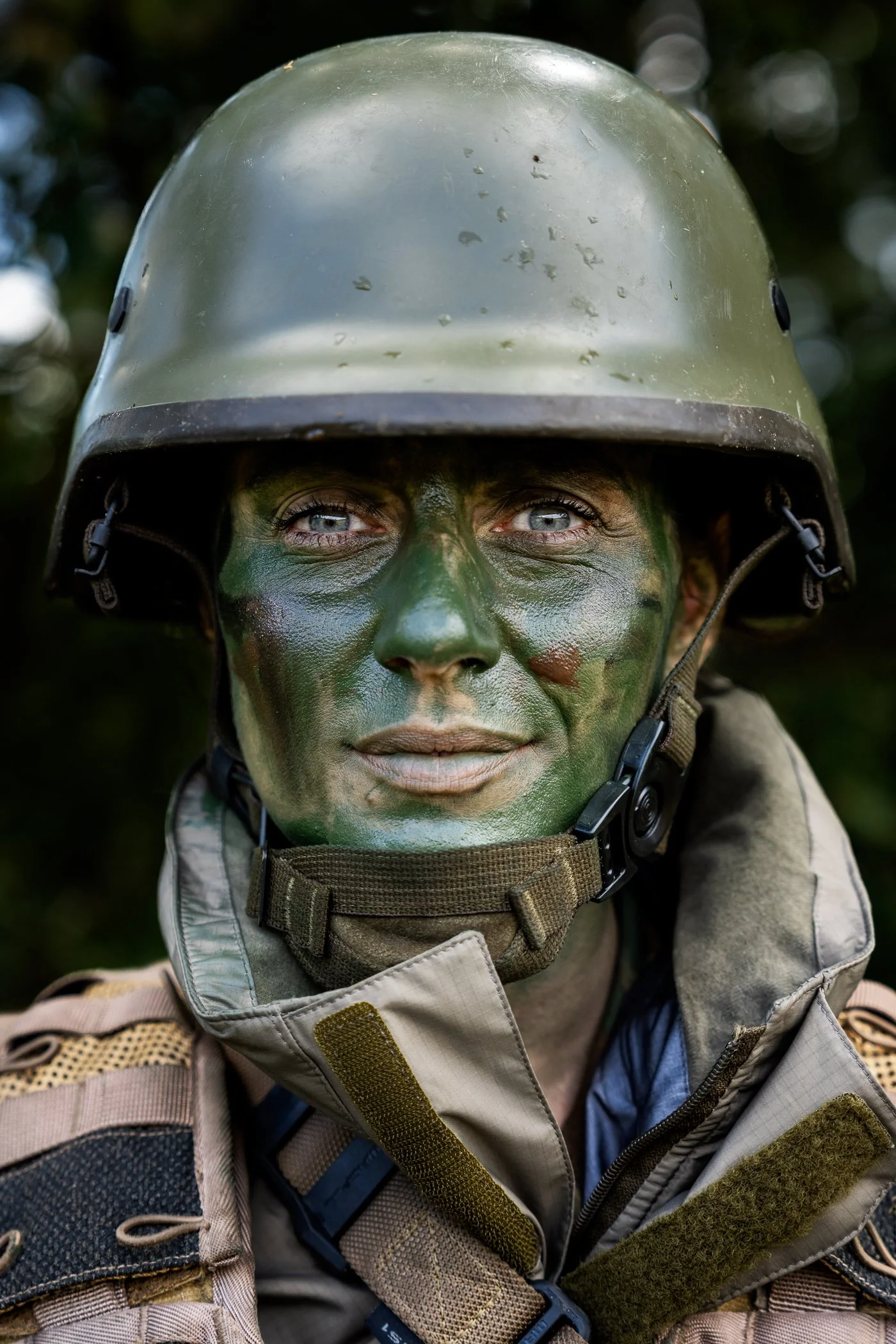Close-up van een militair met gezichtsmake-up en een helm, gekleed in camouflage-uniform, tegen een natuurlijke achtergrond.