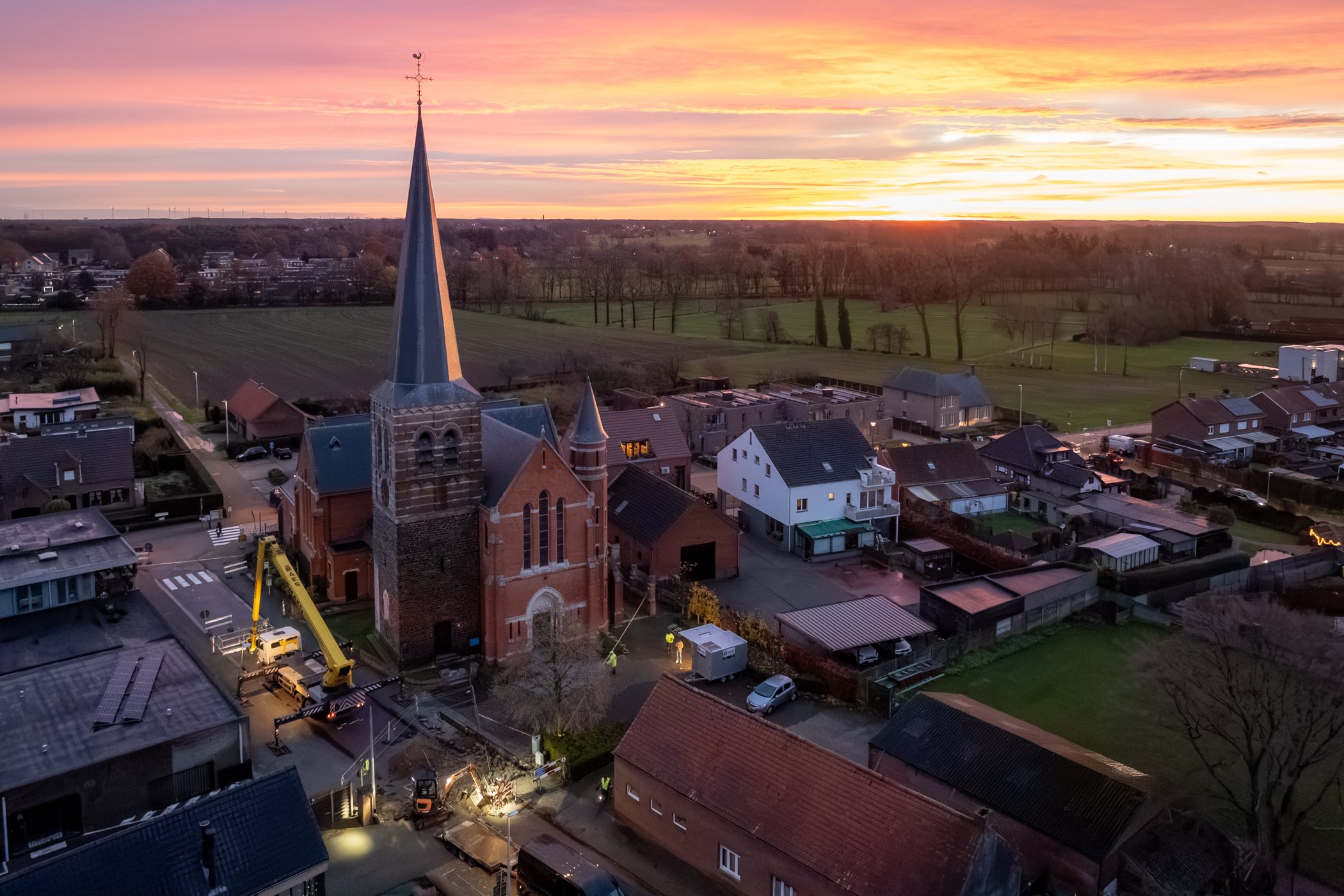 Uitzicht op een kerkje met hoge spits, omringd door huizen en velden bij zonsondergang.