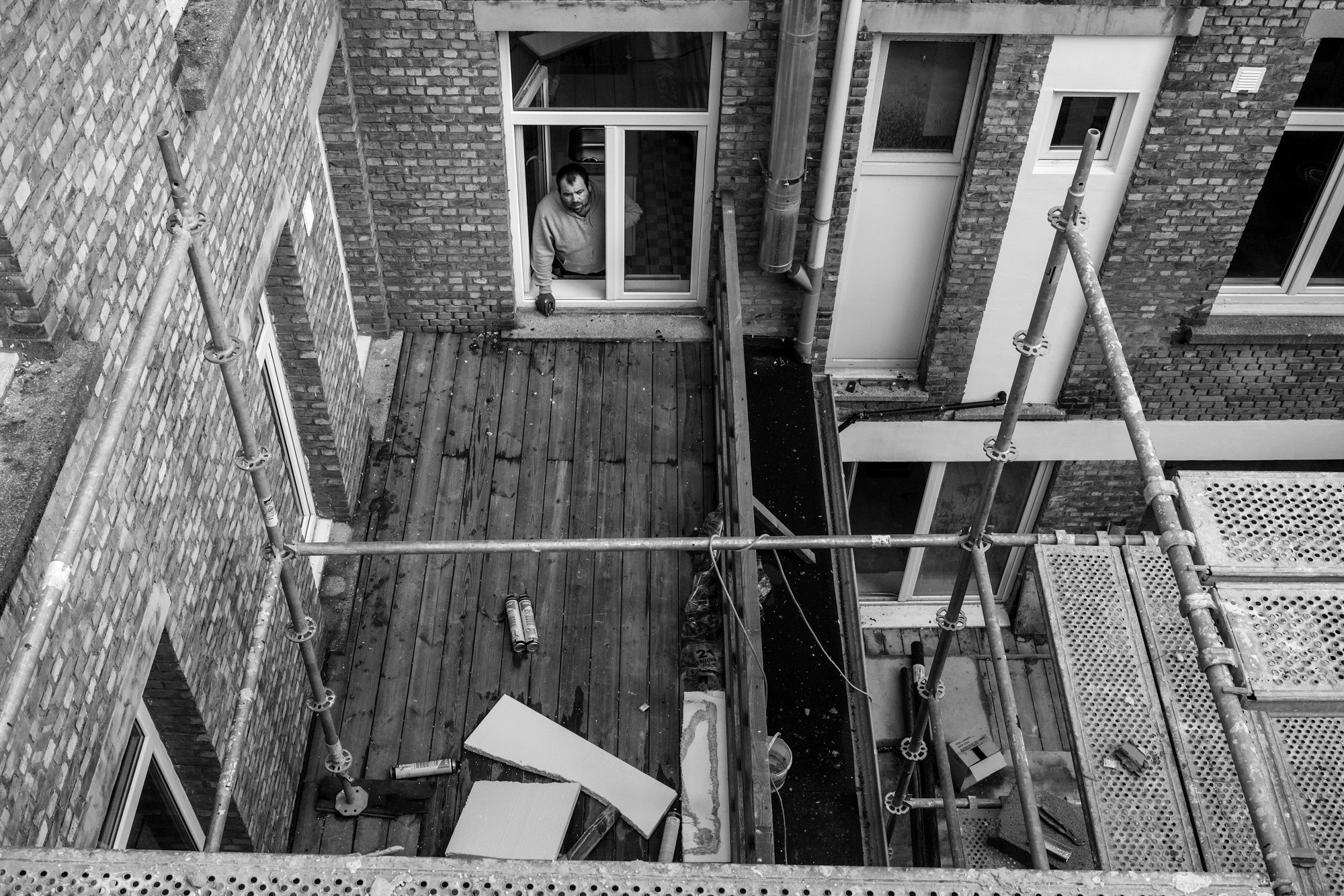 Een man kijkt uit een raam op een bouwplaats met houten vloer op een balkon, omgeven door steiger, met bouwmaterialen en gereedschap.