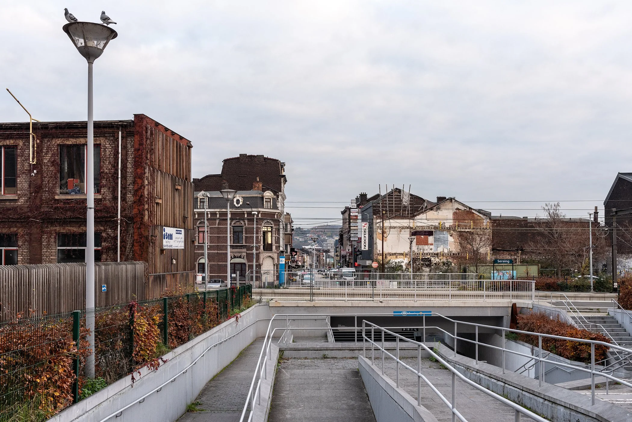 Stadsplein met oude en moderne gebouwen, een straat met verkeer en een metro-ingang in de voorgrond, en een bewolkte hemel.