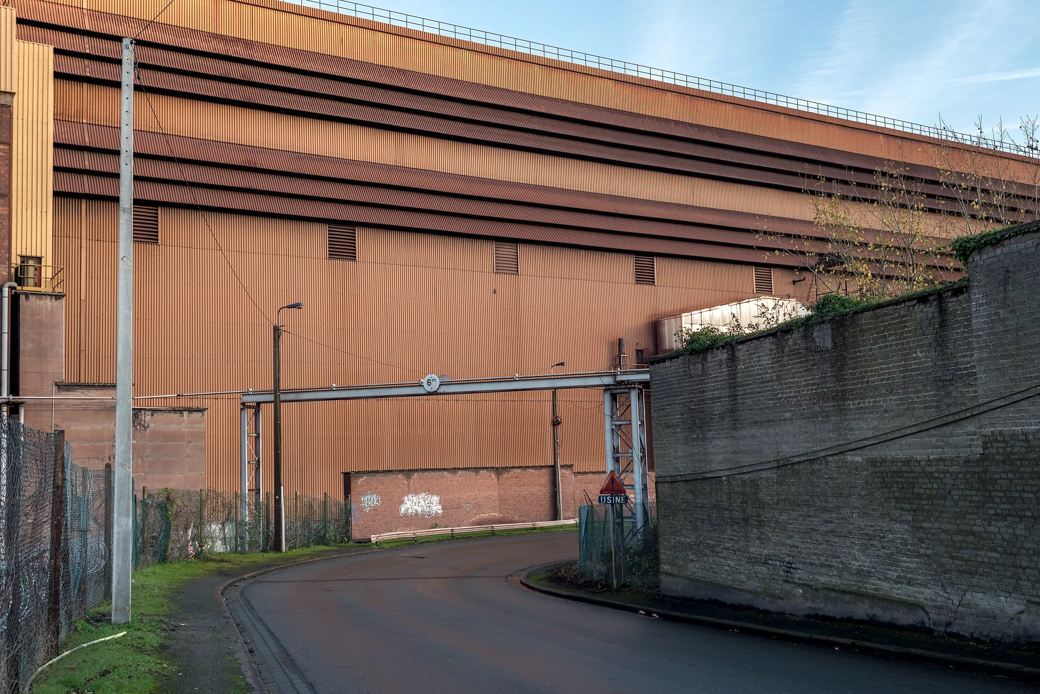 Een weg buigt af links met een hoge, grote oranje loods op de achtergrond. Er is graffiti op een muur en een signaalbord met 'USINE'.