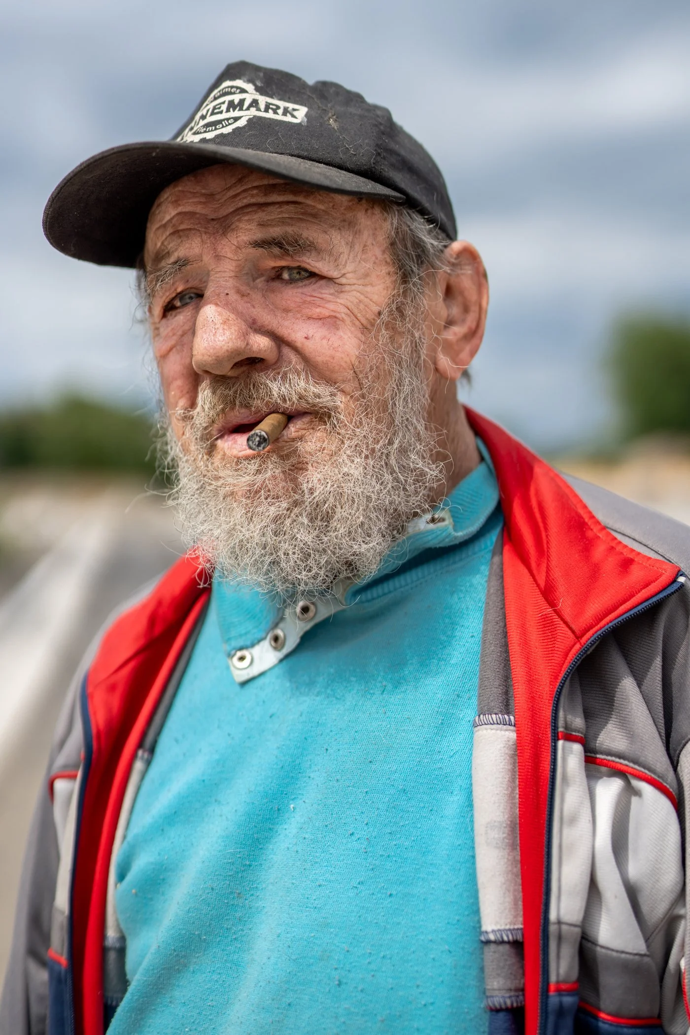 Een oudere man met een grijze baard en een sigaret in zijn mond, draagt een zwarte baseballpet en een kleurrijke jas, buiten op een bewolkte dag.