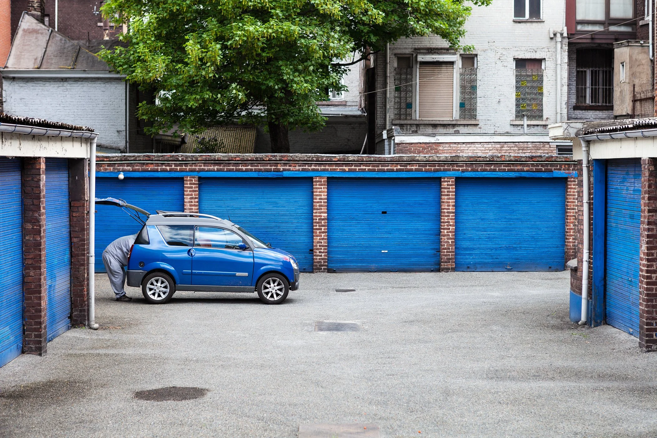Een pleinarena met blauwe garageboxen, een blauwe kleine auto en een persoon die de achterklep opent. Achter de garageboxen staat een boom en een gebouw met ramen.