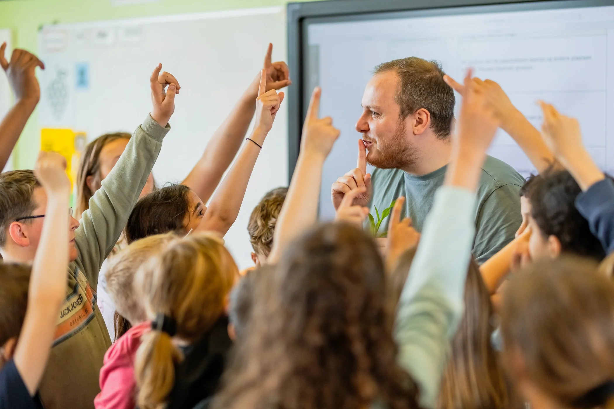 Een man onderwijst een klas vol kinderen die enthousiast hun handen omhoog steken en een vraag willen stellen in een klaslokaal.