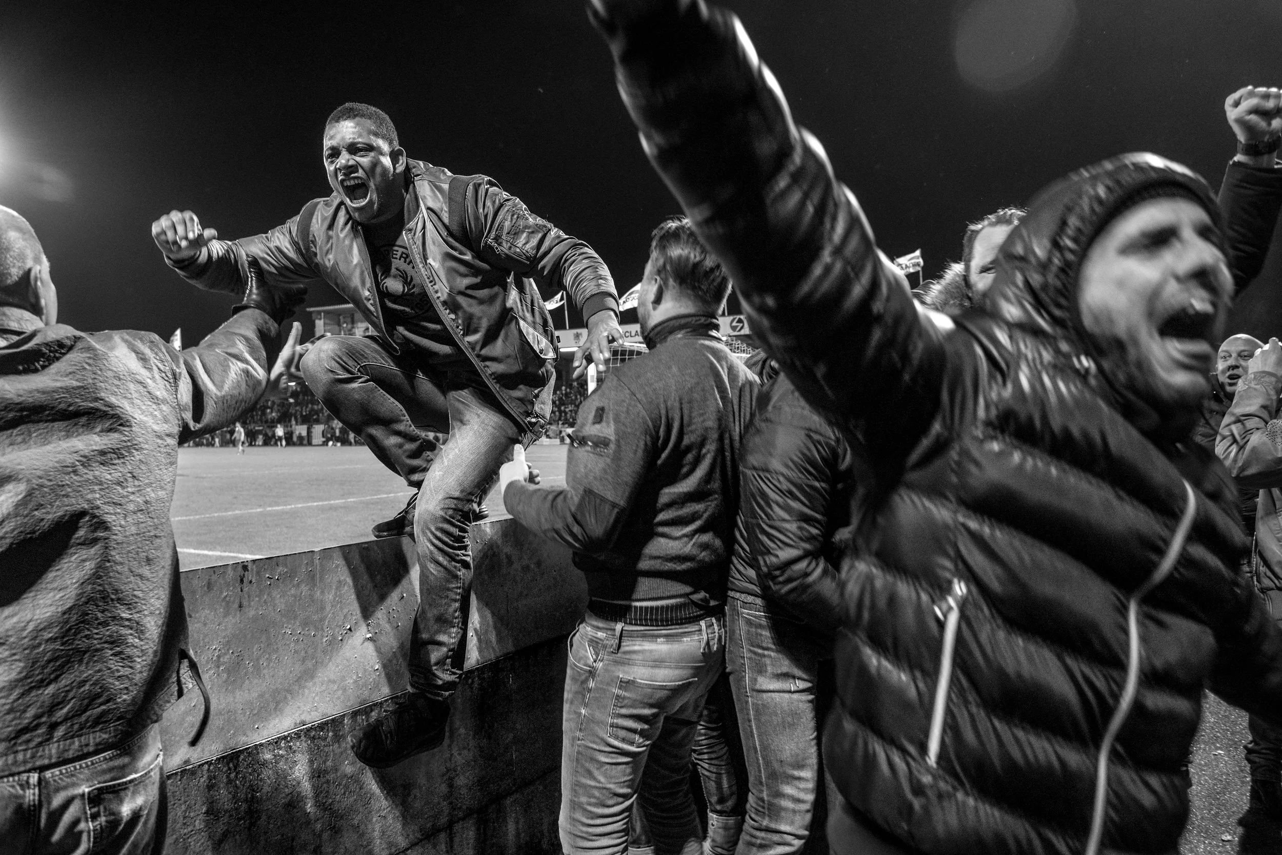Mensen vieren op een sportveld 's nachts, met één persoon die op de rand van de muur springt en andere die juichen en hun vuisten omhoog steken.