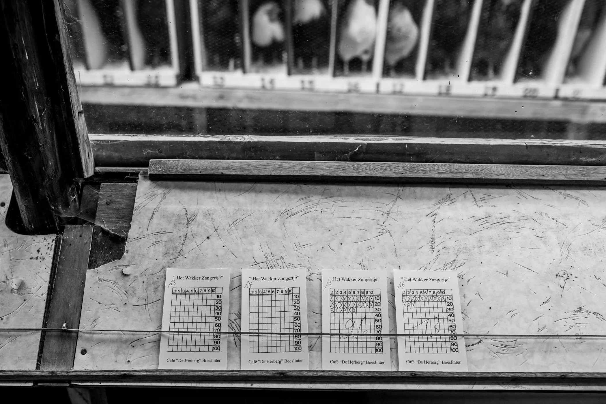 Vier bingo-kaarten liggen op een houten tafel, met een muur en een raam met roosters op de achtergrond.