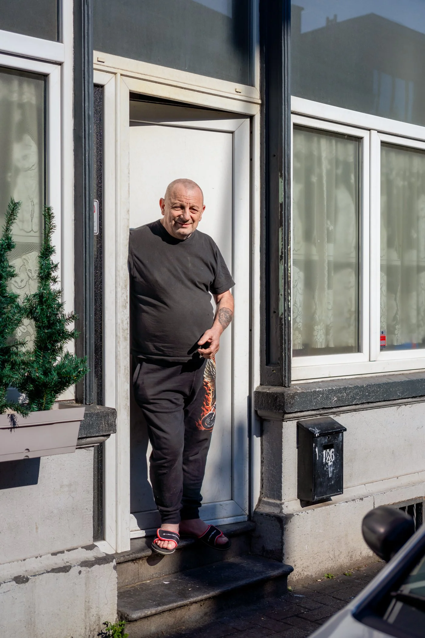 Man staat op de deuropening van een huis, draagt zwarte kleding en slippers, en kijkt naar de camera.