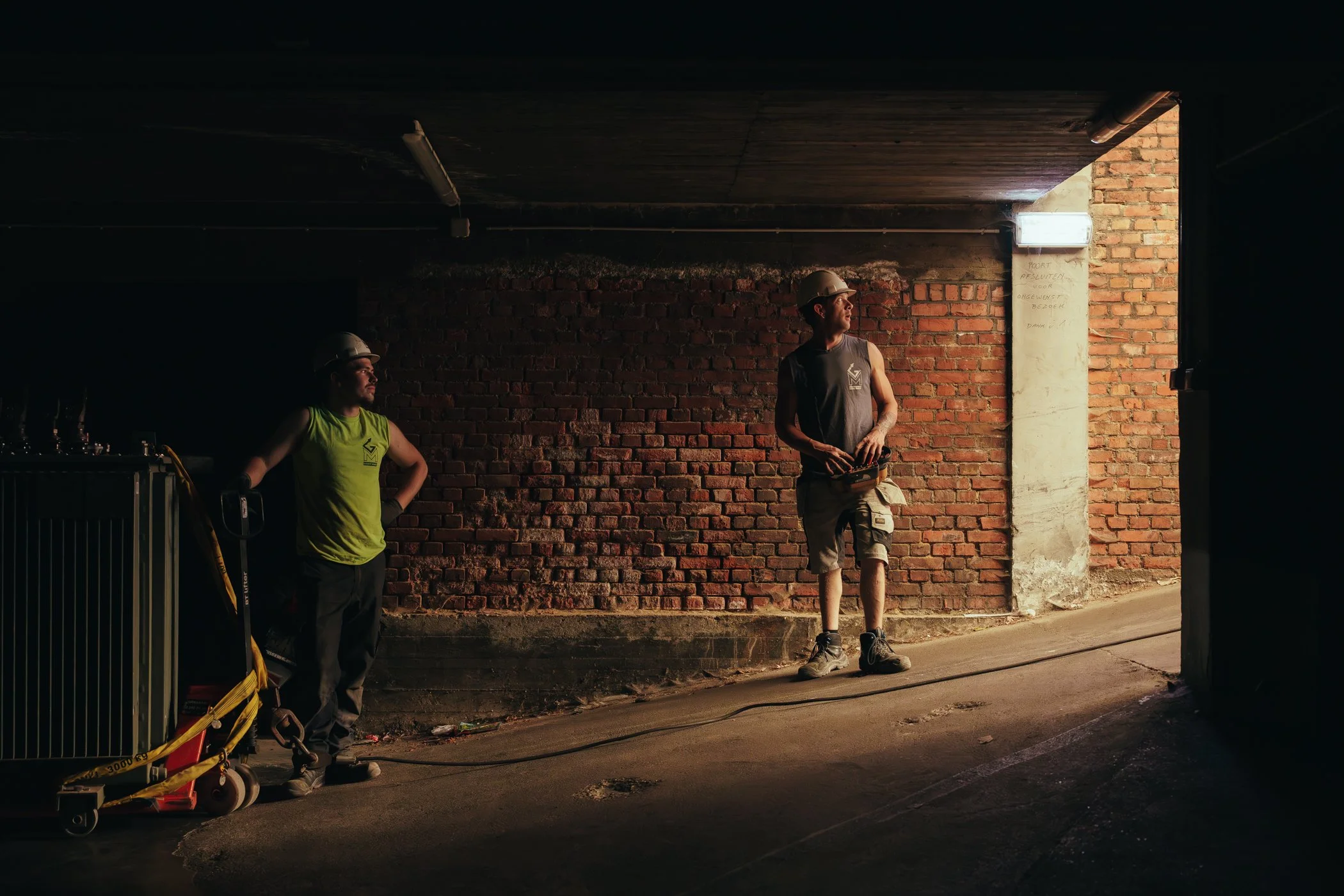 Twee bouwvakkers in een ondergrondse parkeergarage, één leunt tegen de muur terwijl de ander een afstandsbediening vasthoudt.