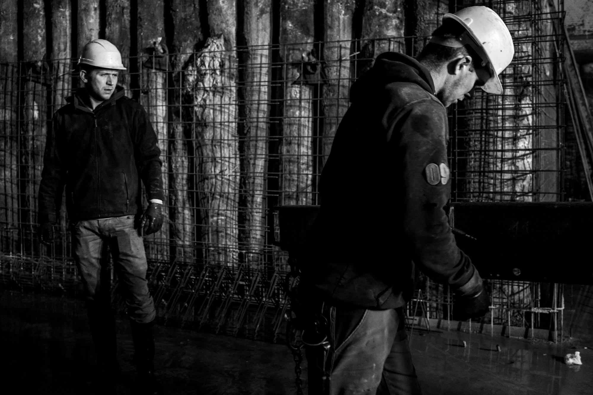 Twee bouwvakkers dragen werkhelmen en werkkleding op een bouwplaats, met staalarmen en gereedschap.