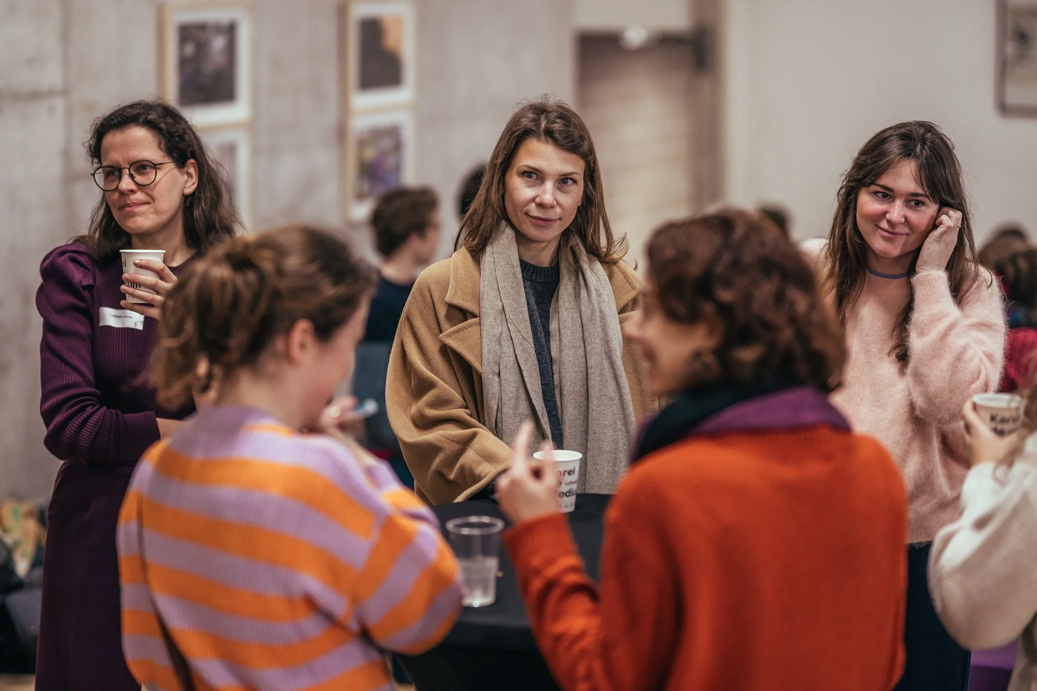 Groep vrouwen die praten en drinken in een informele setting, binnen, decor met foto's op de muur.