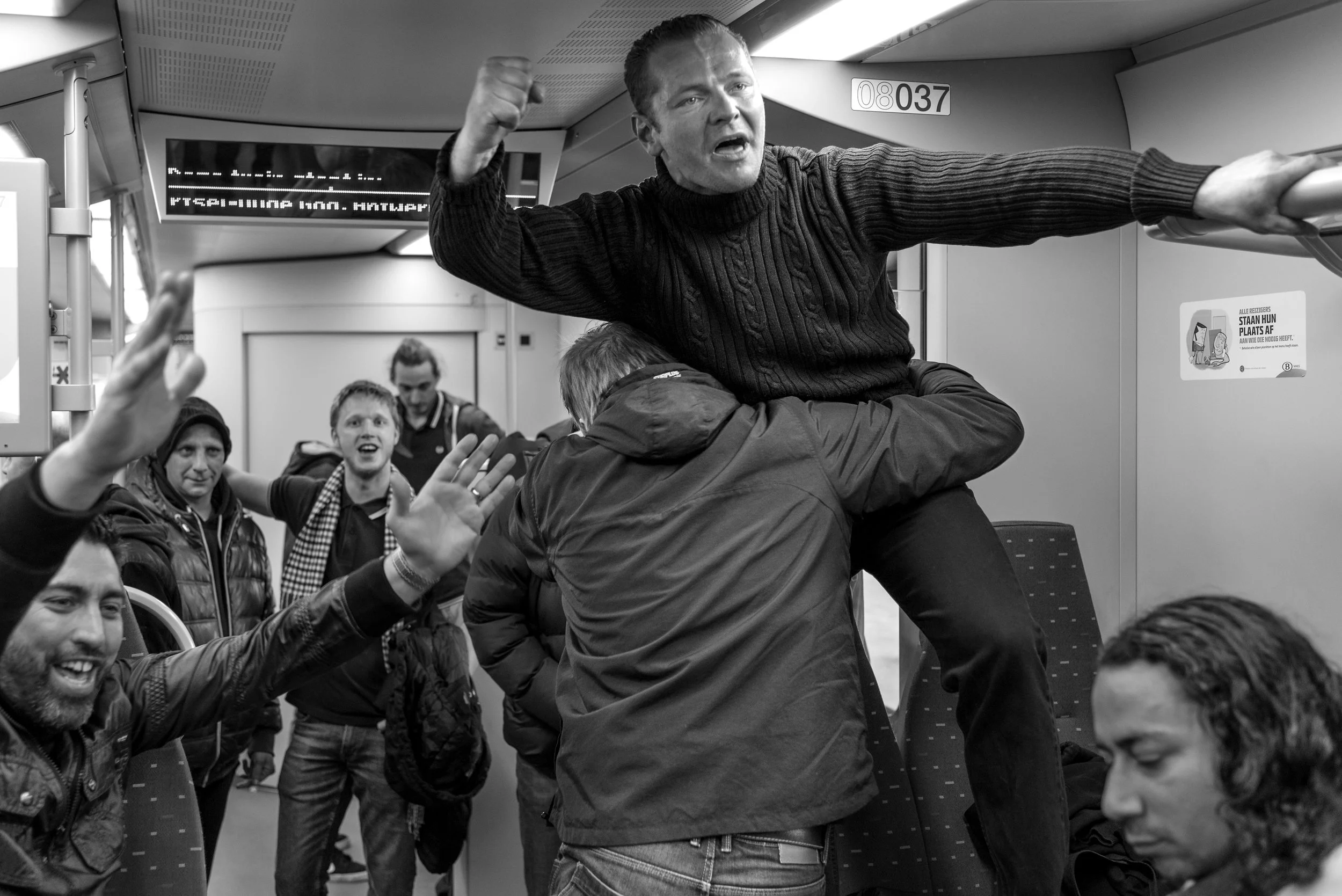 Een groep mensen op een trein, met één persoon die op schouders staat en zich boos uitdrukt, terwijl anderen om hem heen reageren.