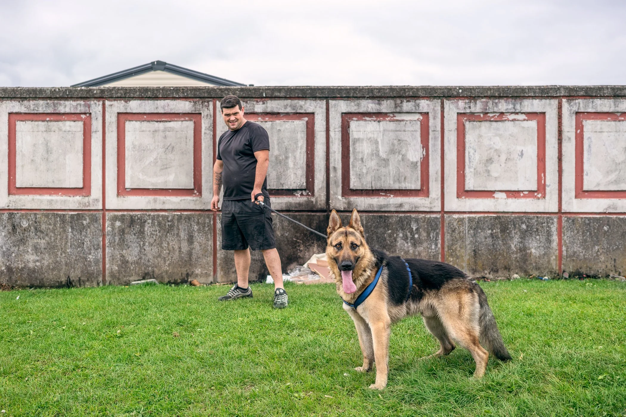 Man met een grote Duitse herdershond op een grasveld, met een betonnen en rode muur op de achtergrond en een lucht met bewolking