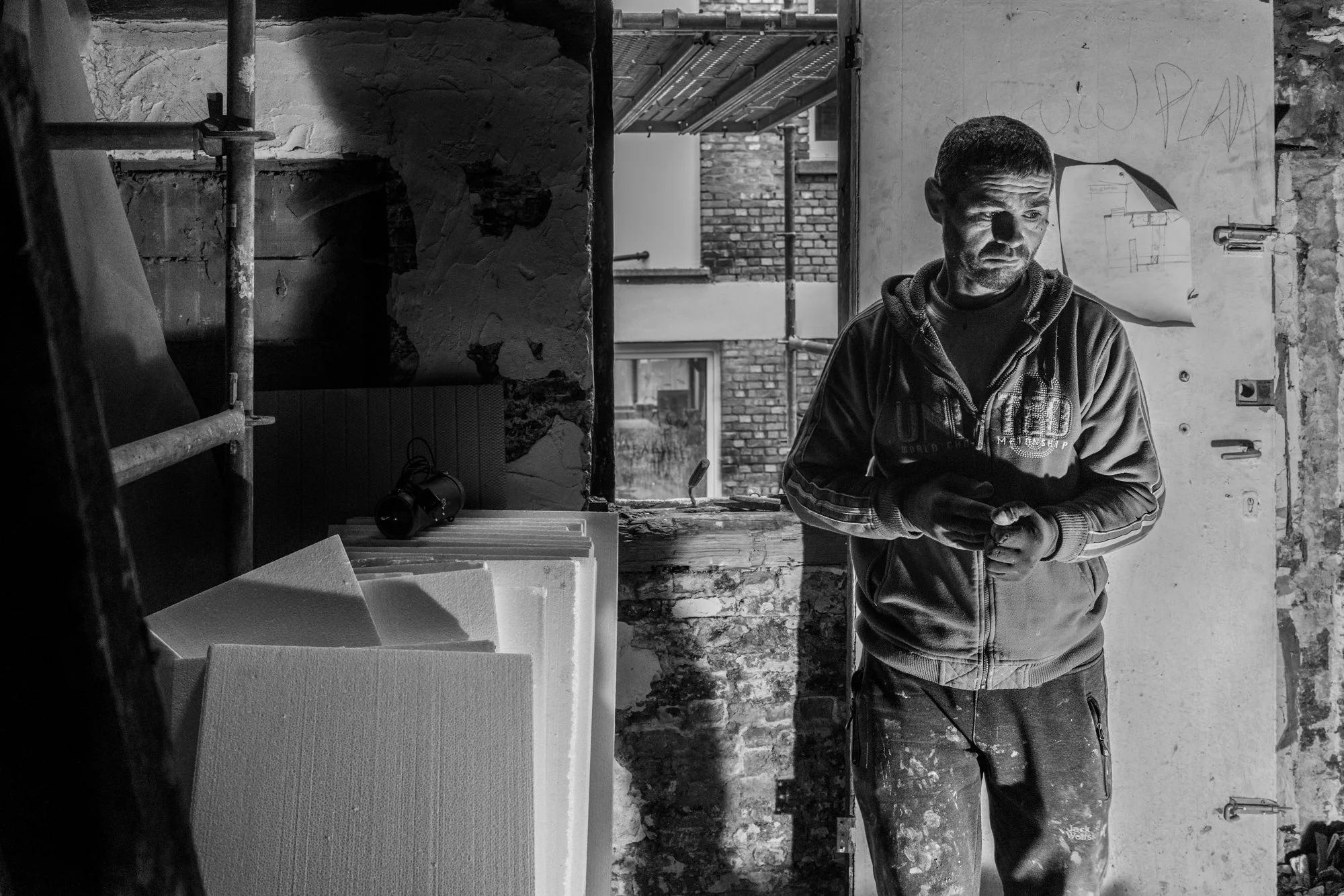 Man in een bouwplaats met oude muren en raam, hij kijkt naar een telefoon, omringd door bouwmaterialen en gereedschap, in zwart-wit.