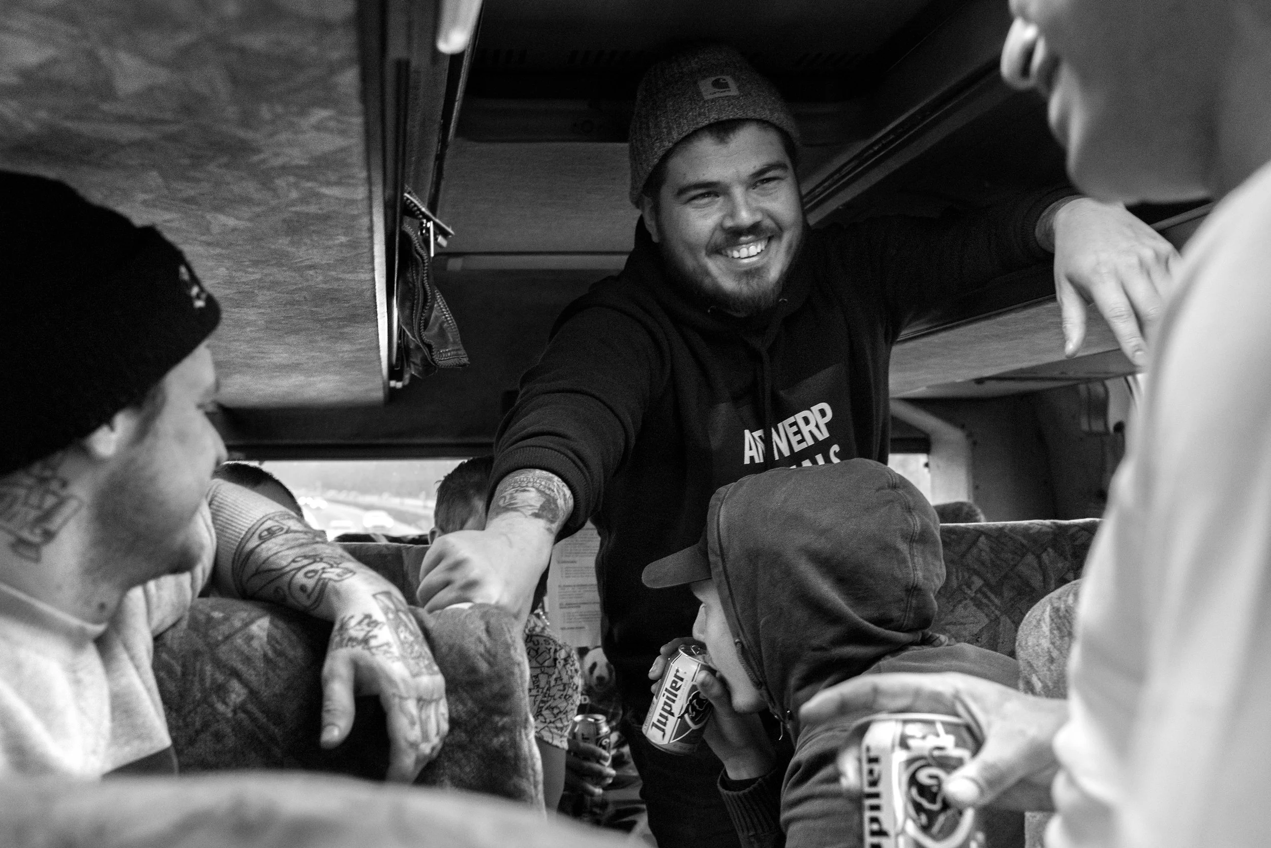 Groep jonge mensen die lachen en praten op een bus, een man met een grijze muts en een tattoo glimlacht naar de camera. Ze drinken en hebben een gezellige sfeer.
