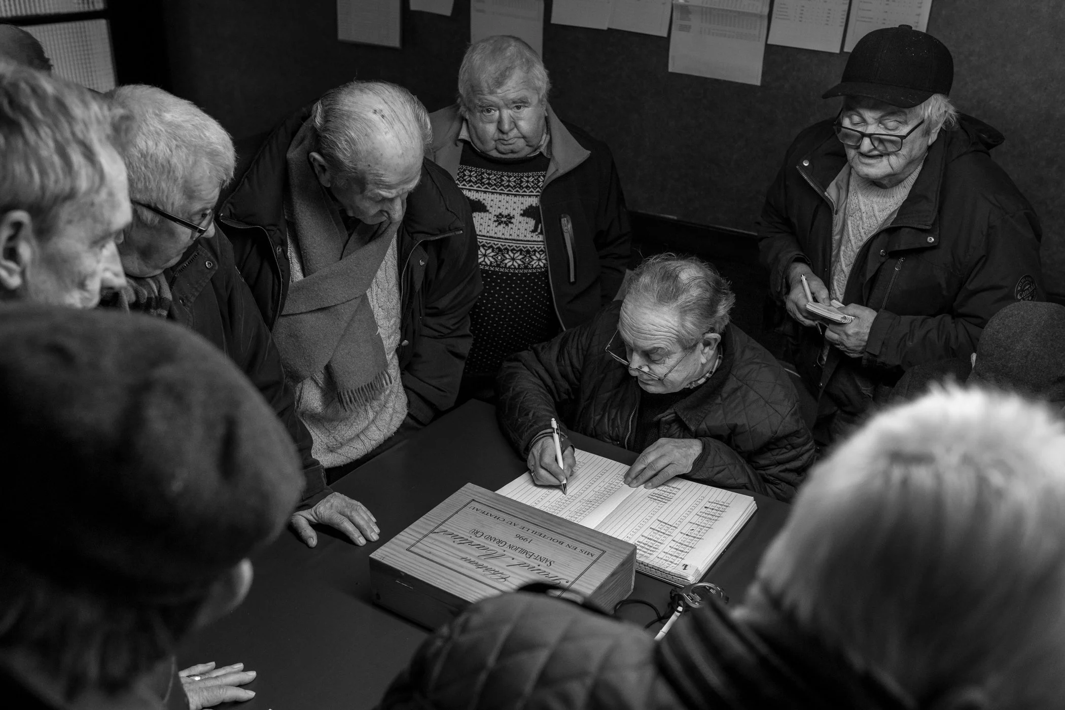 Een groep ouderen verzamelt zich rond een man die een inschrijving bijhoudt op een formulier op een tafel, in een zwart-wit foto.