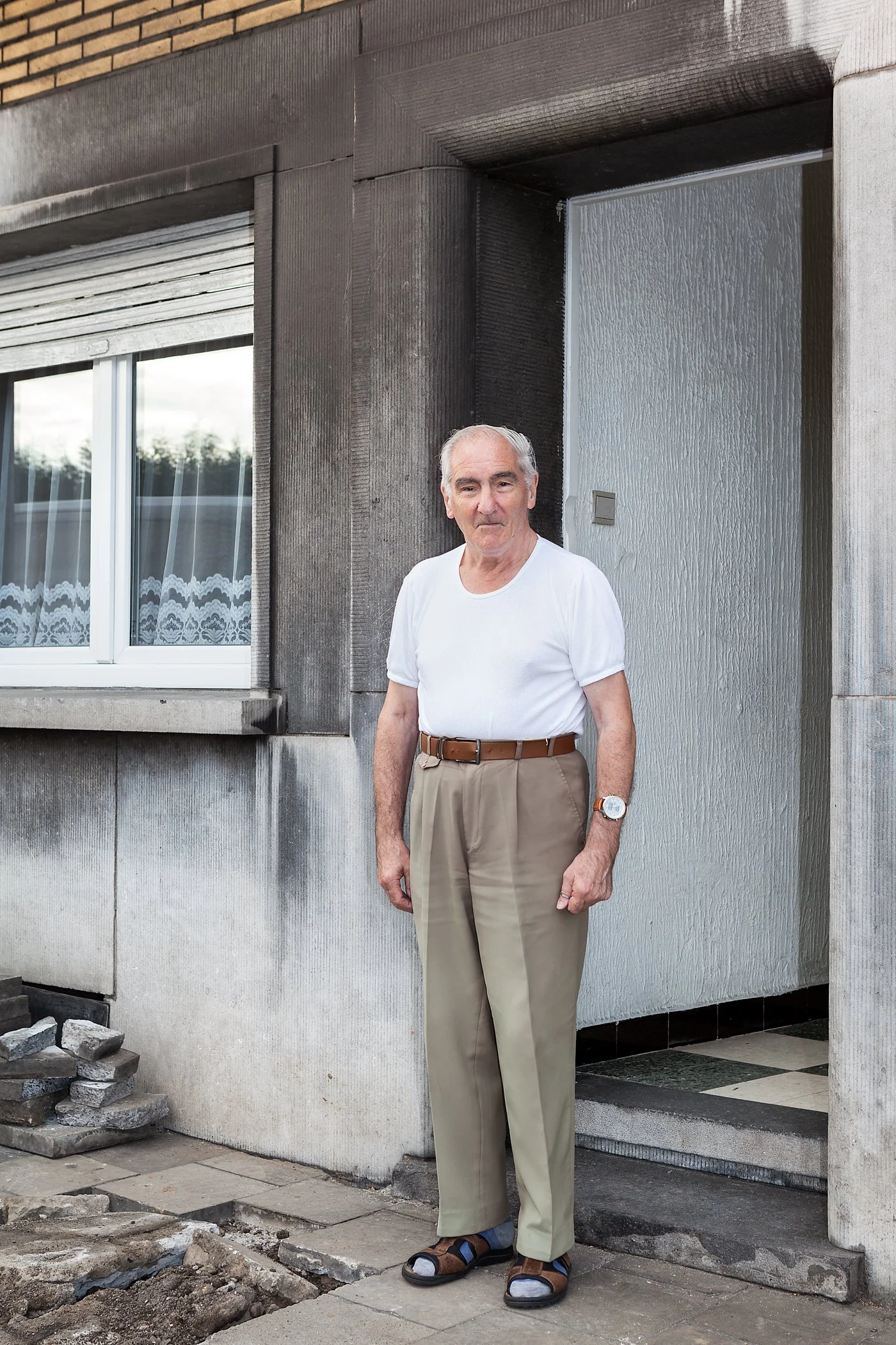Oudere man staat buiten voor een huis met een grijze gevel, zwarte raamkozijnen en een deur, draagt een witte t-shirt, beige broek, sandaal en een horloge.