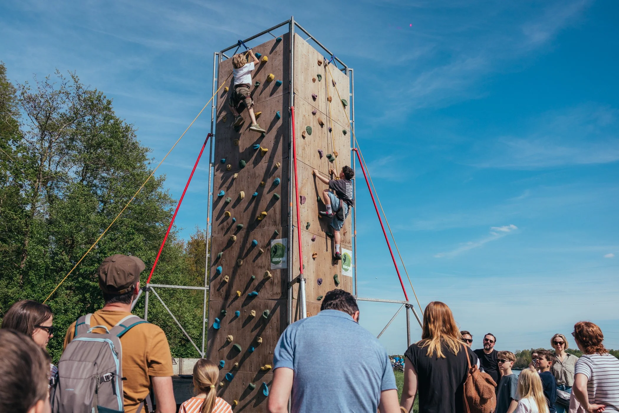 Kinderen klimmen op een grote klimwand tijdens een buitenevenement op een heldere dag, met een groep mensen die toekijken.