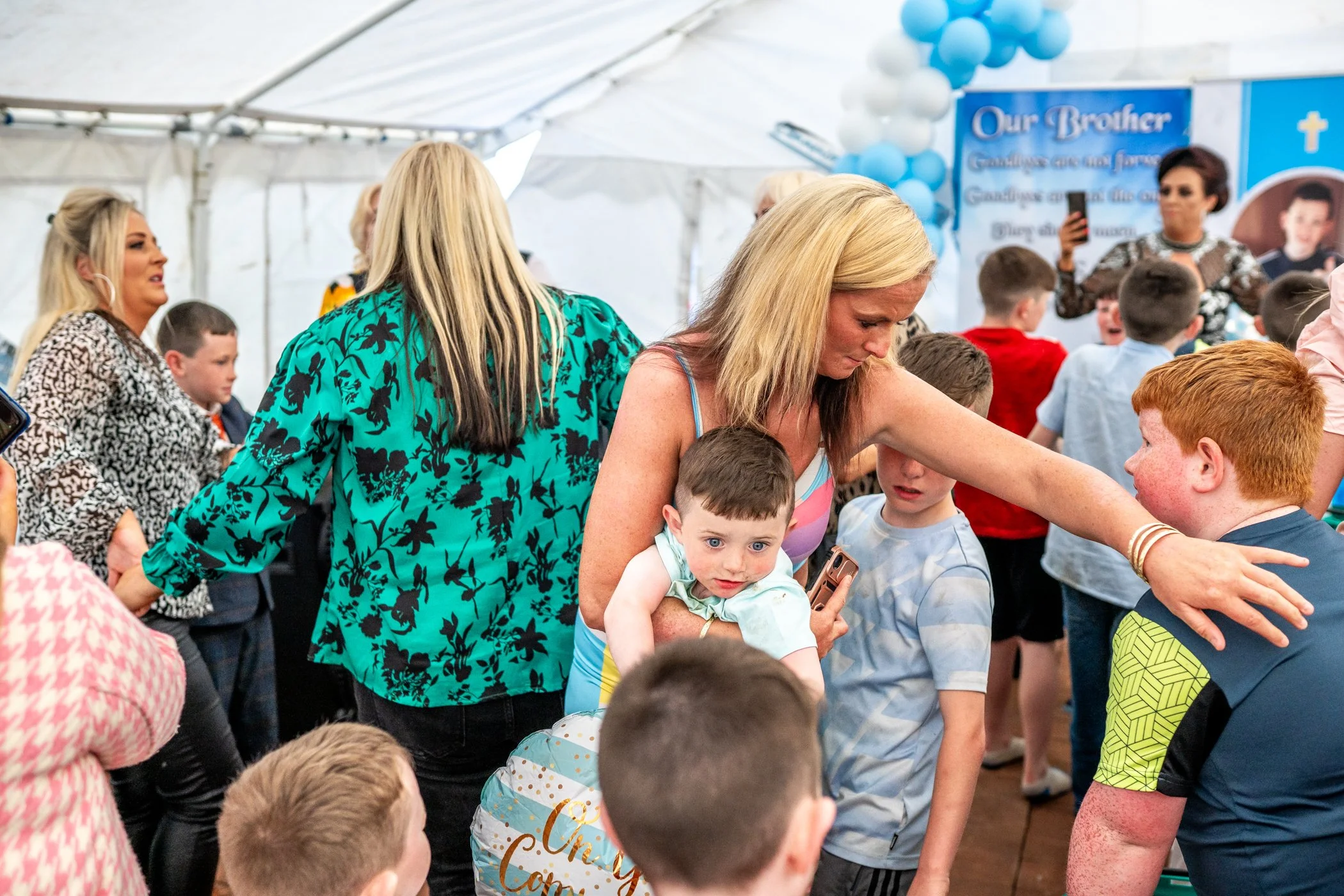 Een verzameling mensen, waaronder veel kinderen, in een witte tent met blauwe ballonnen en een blauw achtergrond, mogelijk tijdens een feest of gemeenschapsevenement.