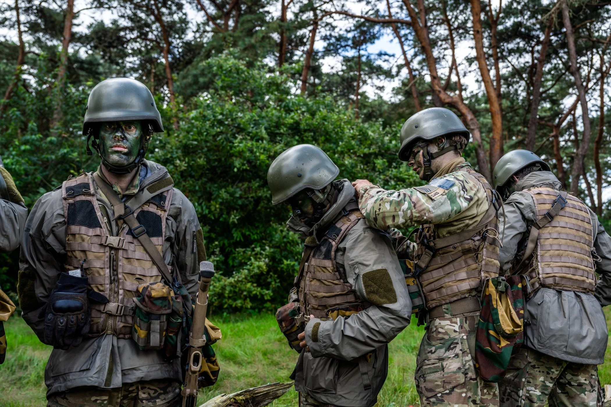 Vier militairen in camouflageuniform en helmen, met uitrusting, staan in een bos, één helpt een ander met uitrusting.
