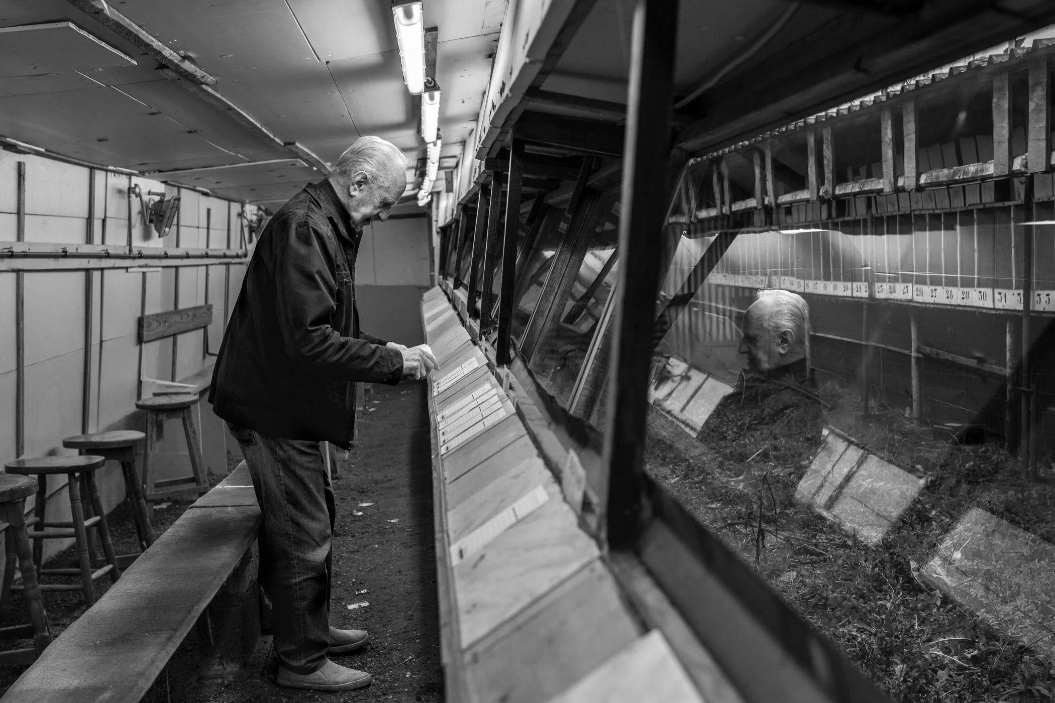 Een oudere man met wit haar en een donkere jas, die naar een vitrinekast met watertanks kijkt in een donkere ruimte, met zijn reflectie zichtbaar in de glasschermen.