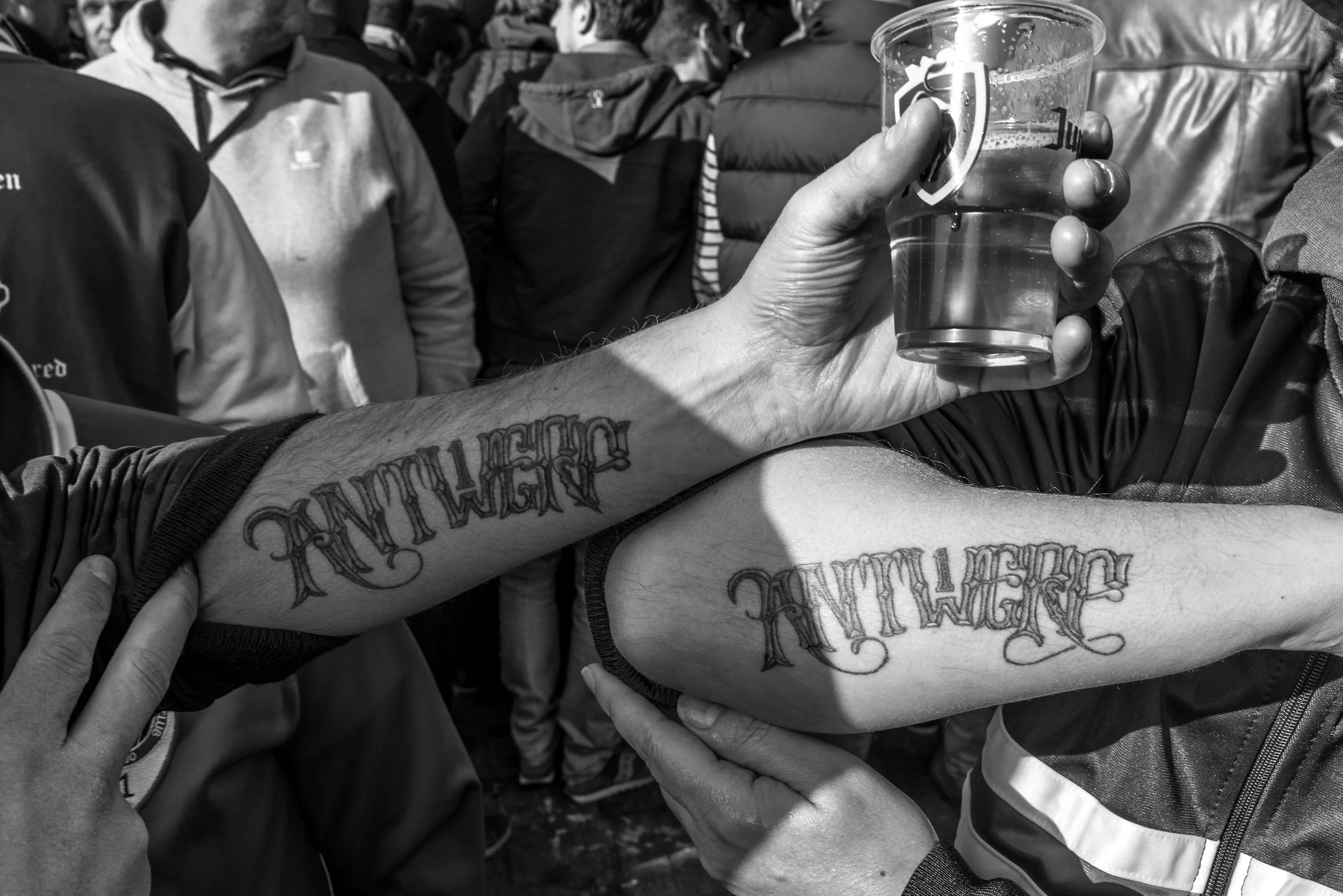 Twee mensen tonen hun getatoeëerde armen met het woord 'ANTWEN' in een gothische stijl, iemand houdt een glas bier vast en er is een groep mensen op de achtergrond.