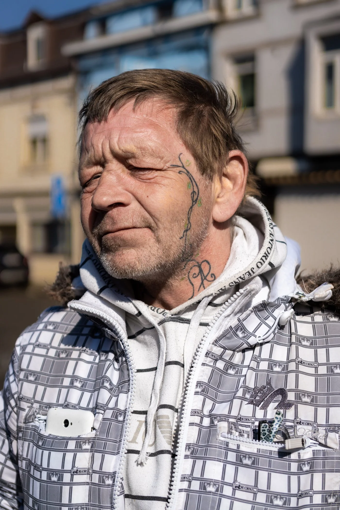 Een man met gezichtstatoeage die in de zon staat, gekleed in een witte gestreepte hoodie en een witte jas met patronen, op een straat met gebouwen op de achtergrond.