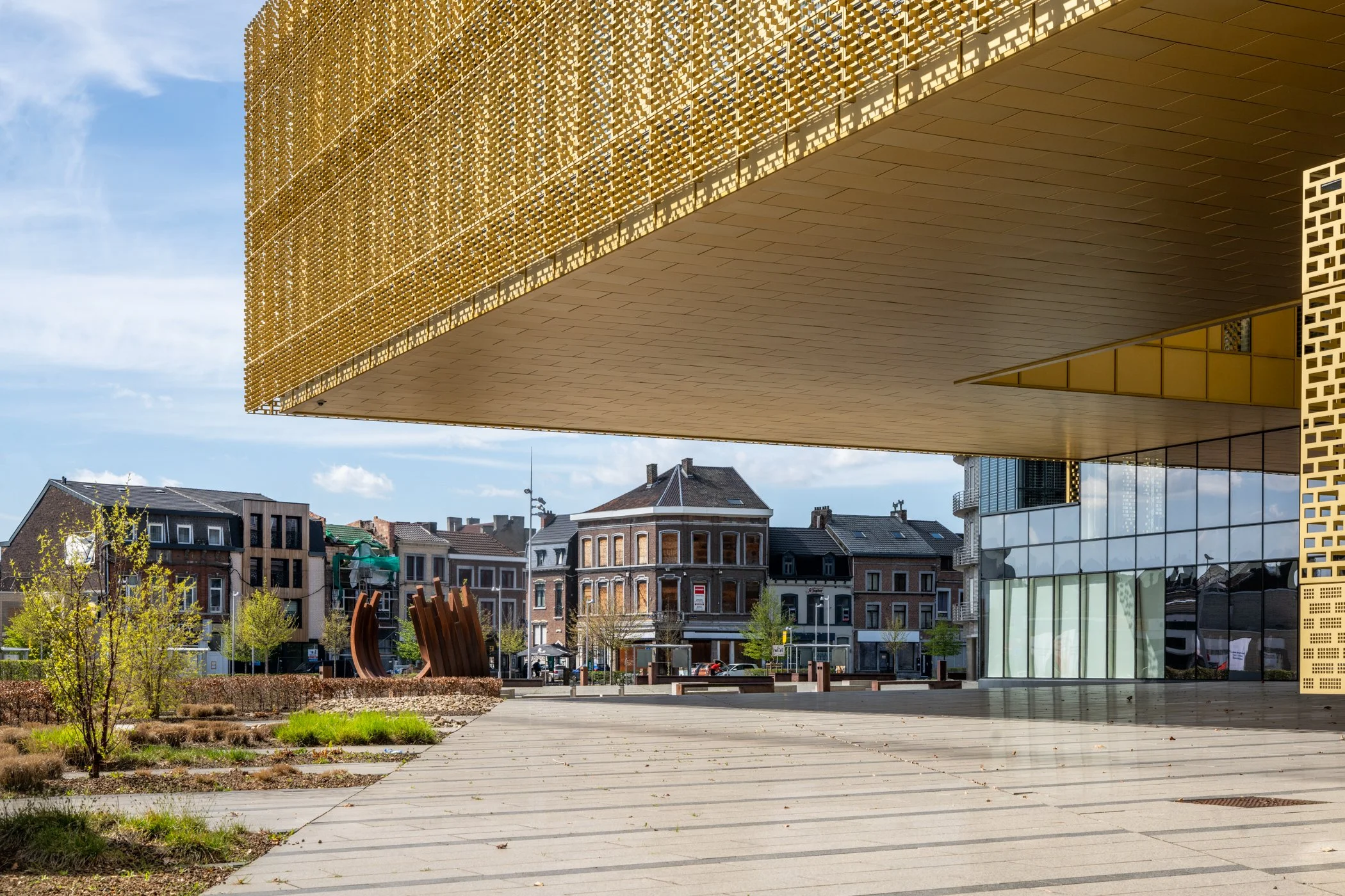 Moderne gebouwen met een grote overkapping in een stadsplein, met enkele bomen en kunstwerken op de voorgrond, onder een blauwe lucht met wat wolken.